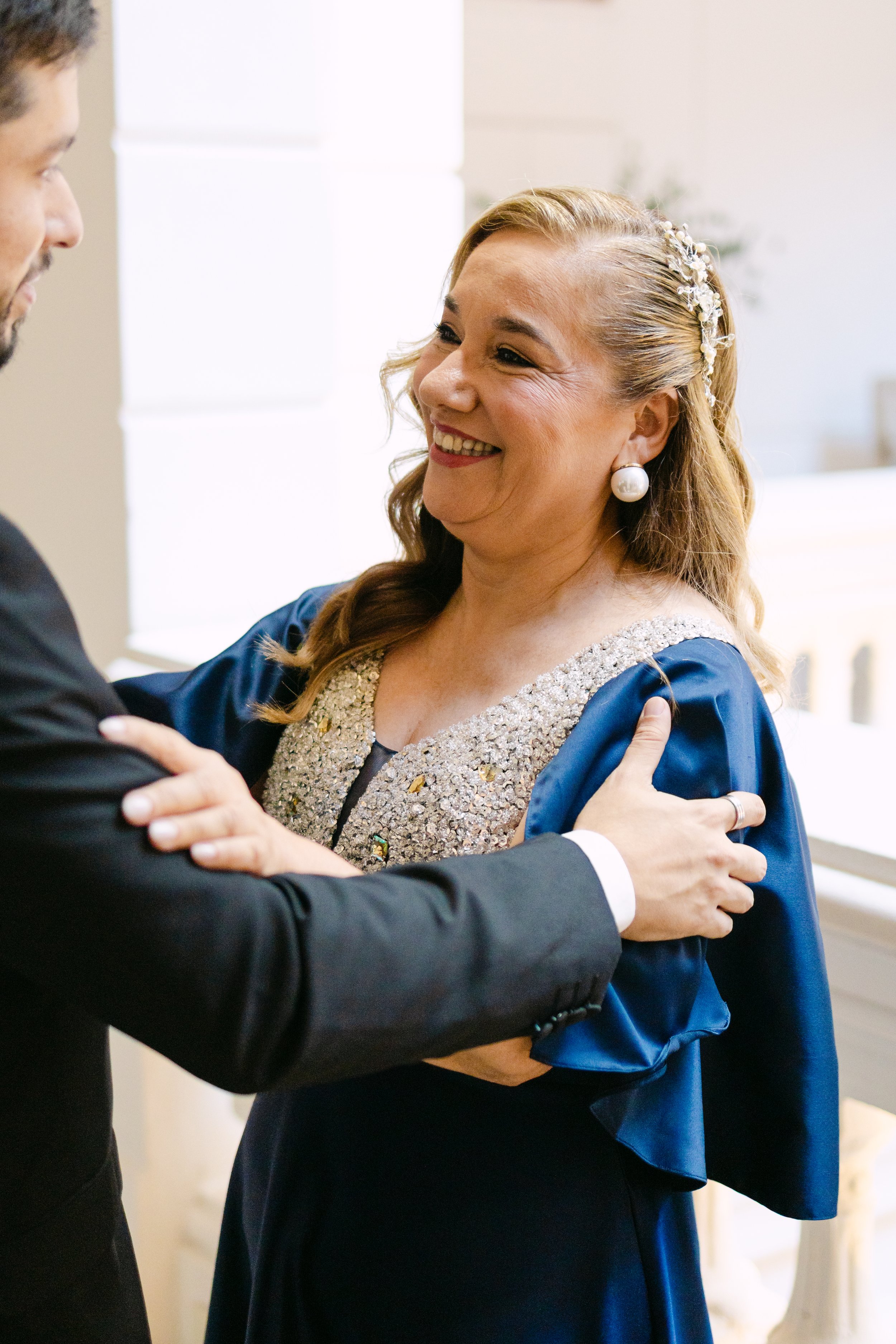 Una mujer sonriendo, vestida con un vestido azul con detalles de pedrería, se le ve un peinado con una diadema decorativa y un pendiente de perla, está abrazando a un hombre que lleva un traje oscuro.