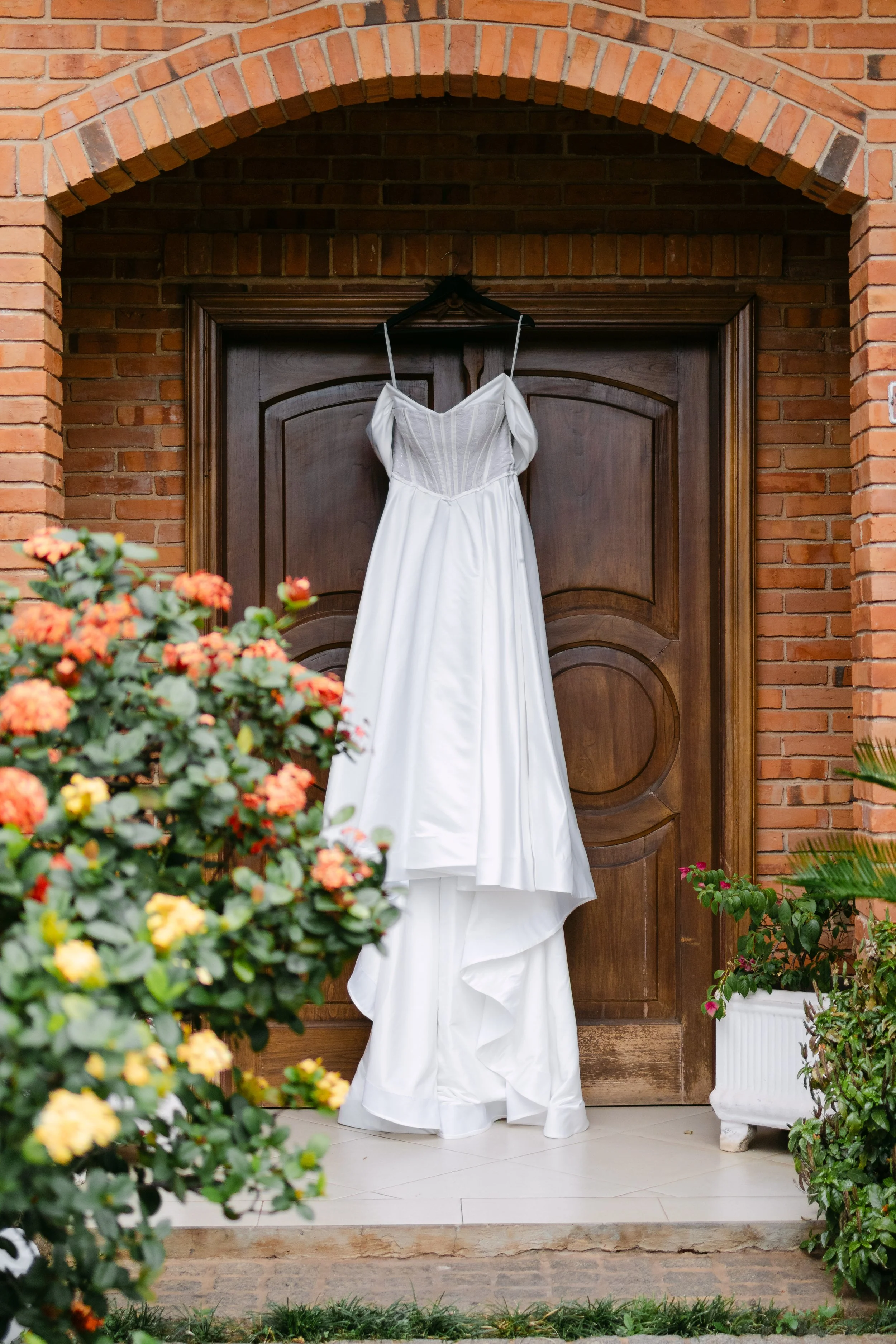 Vestido de novia colgado en una puerta de madera en un fondo de paredes de ladrillo, rodeado de flores de colores en macetas.