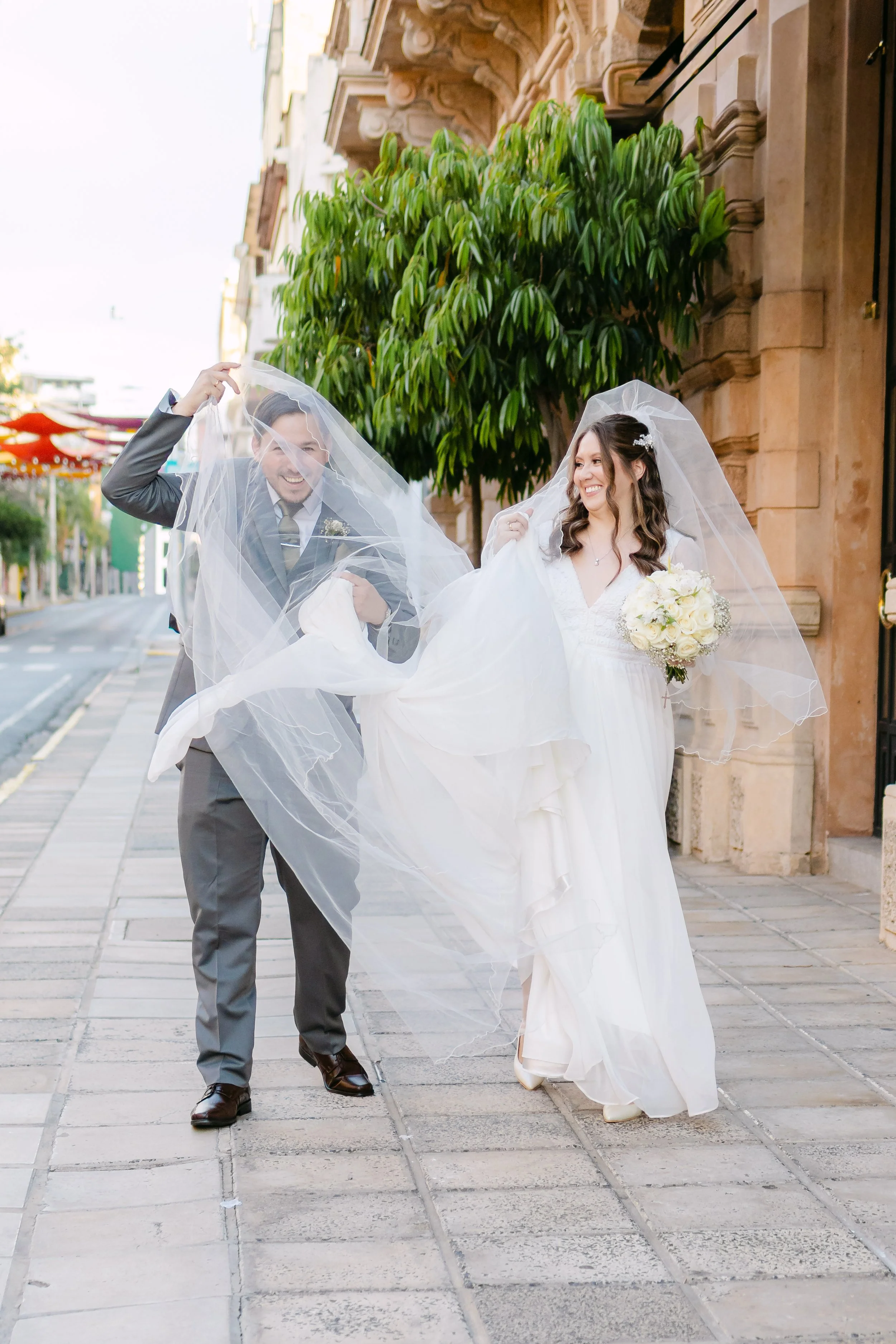 Una pareja de recién casados caminando por la calle, la novia sostiene un ramo de flores blancas y el novio levanta parte de la sombra de la novia, ambos sonrientes bajo un velo de novia. La escena es urbana, con árboles y edificios en el fondo.