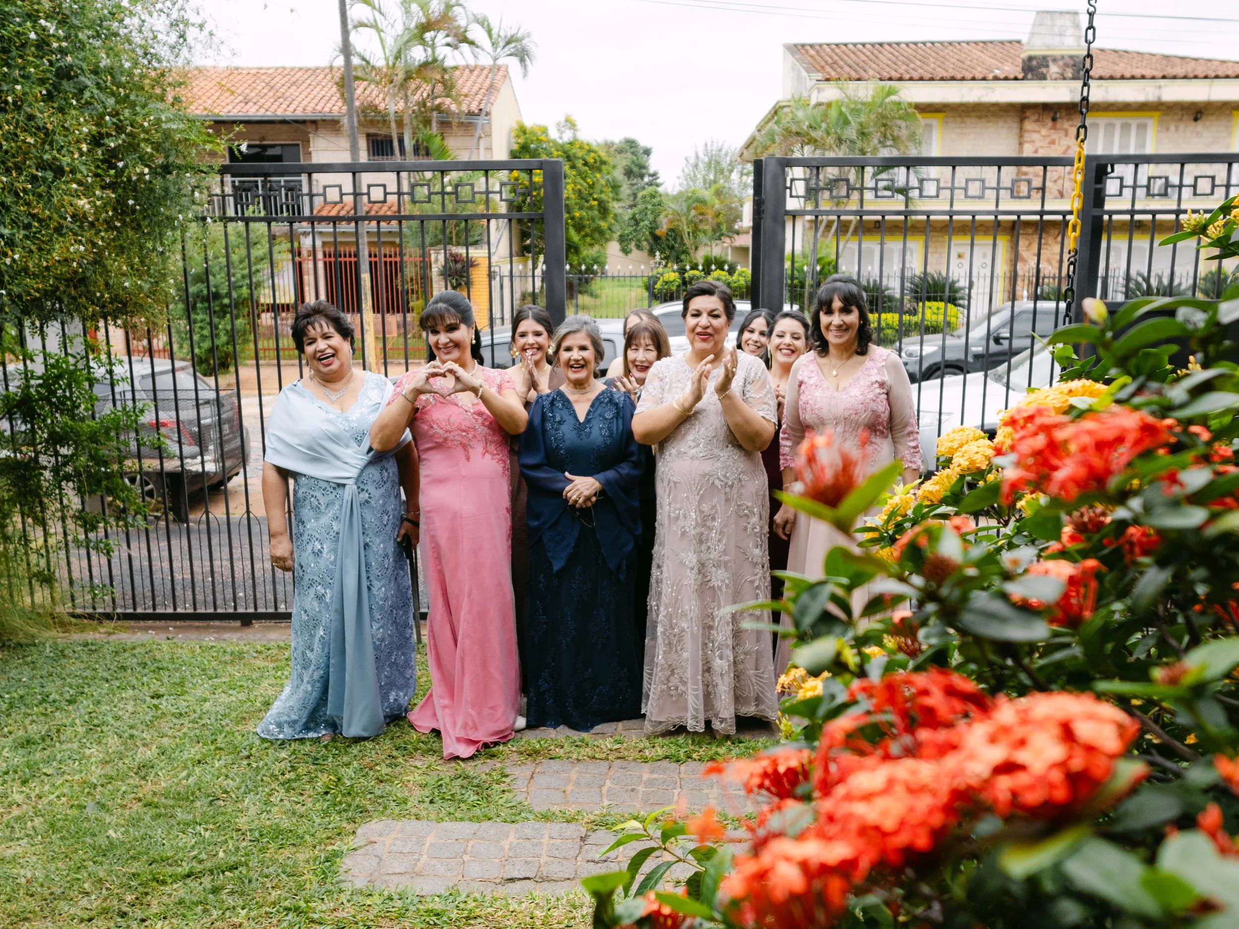 Grupo de mujeres sonriendo y posando delante de una reja de metal, en un jardín con flores de colores y casas de fondo.