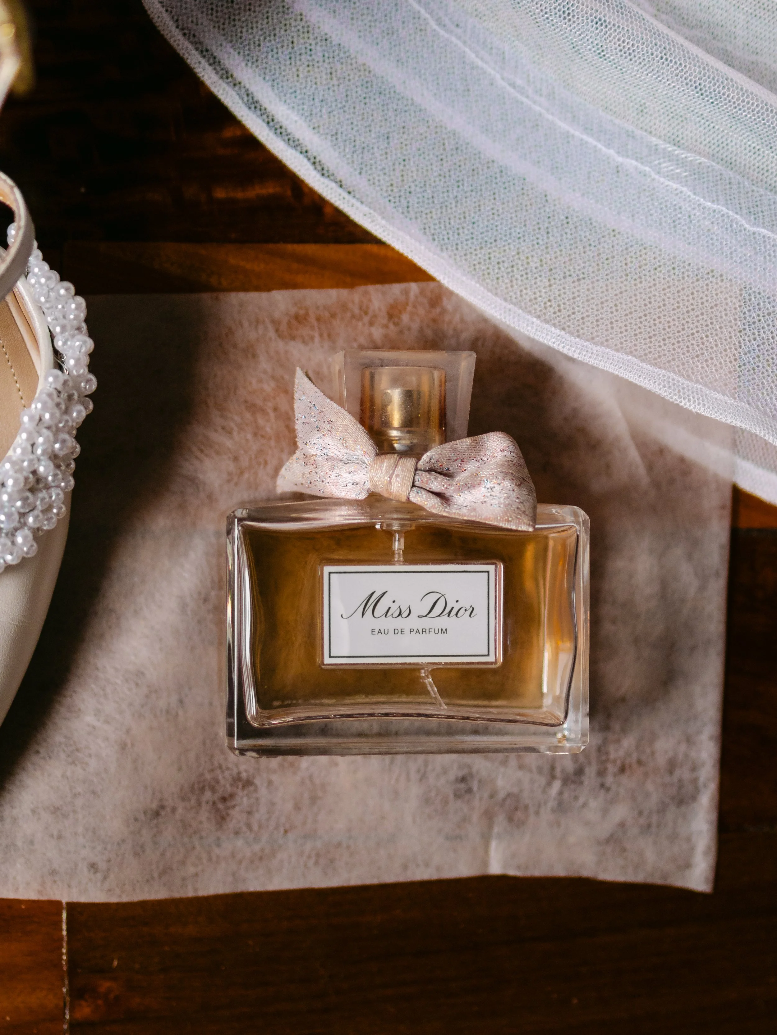 Botella de perfume Miss Dior Eau de Parfum sobre una mesa de madera, con un lazo de tela en la parte superior de la botella y un fondo de tela de encaje blanco.
