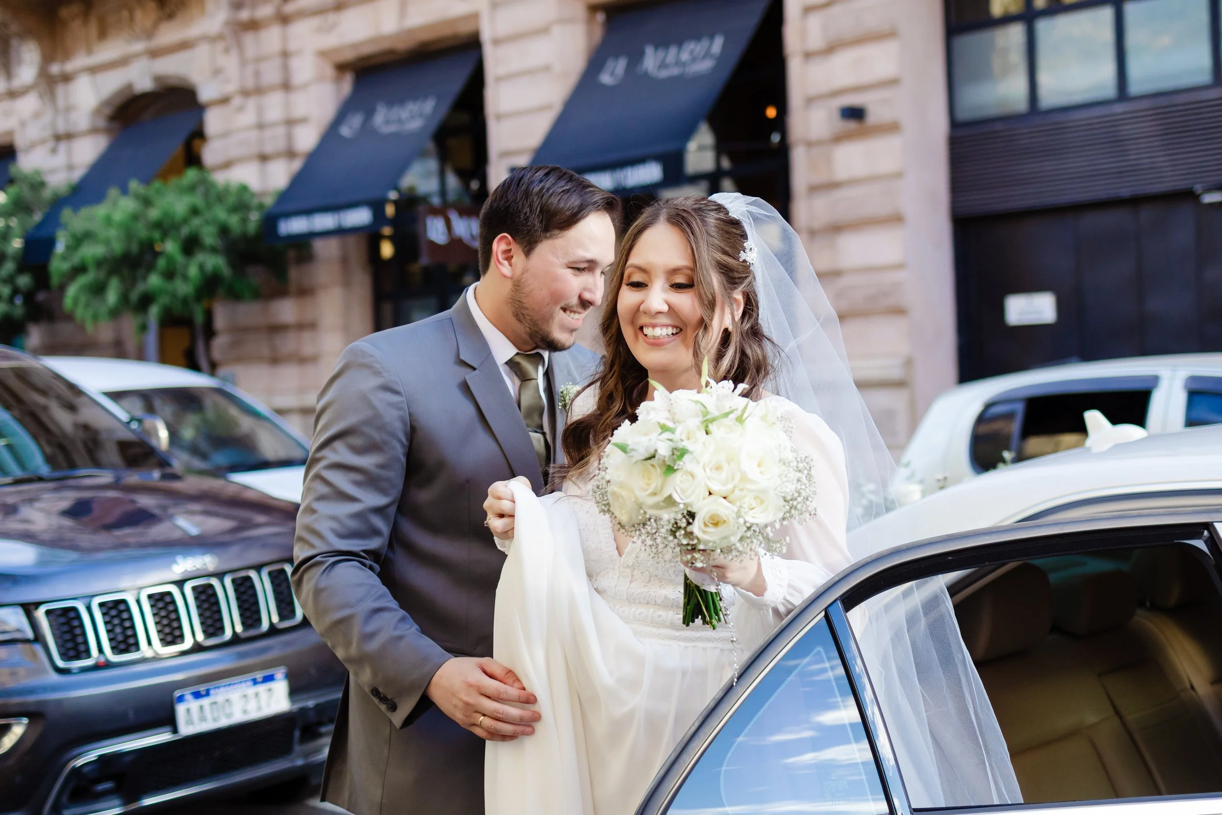 pareja de recién casados en la calle, ella con vestido blanco y ramo de flores, él en traje gris, frente a un auto, sonriéndose, en un entorno urbano con autos y edificio de fondo.