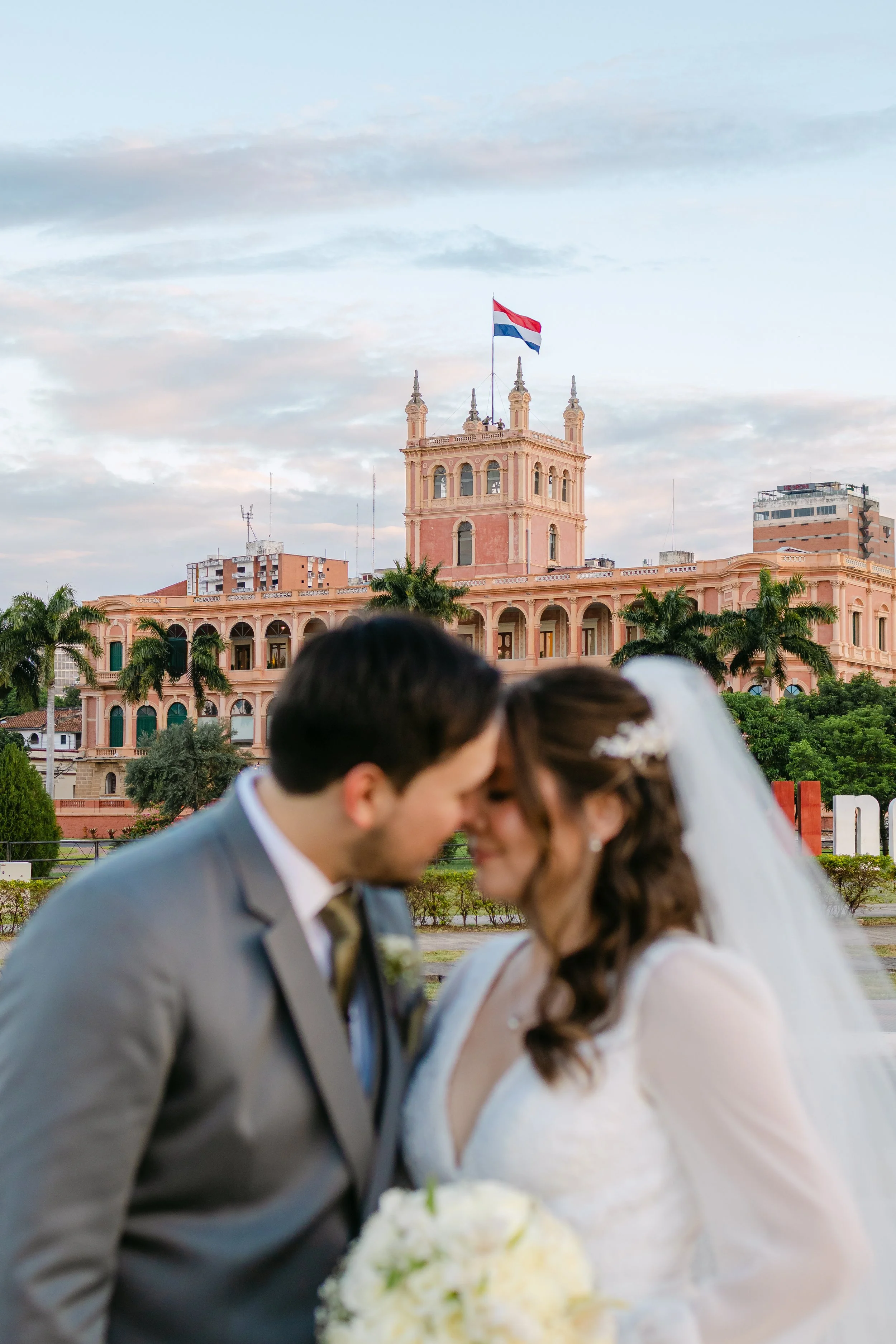 Una pareja de novios en su boda, en primer plano, con un edificio rosa y un árbol con palmas en el fondo.