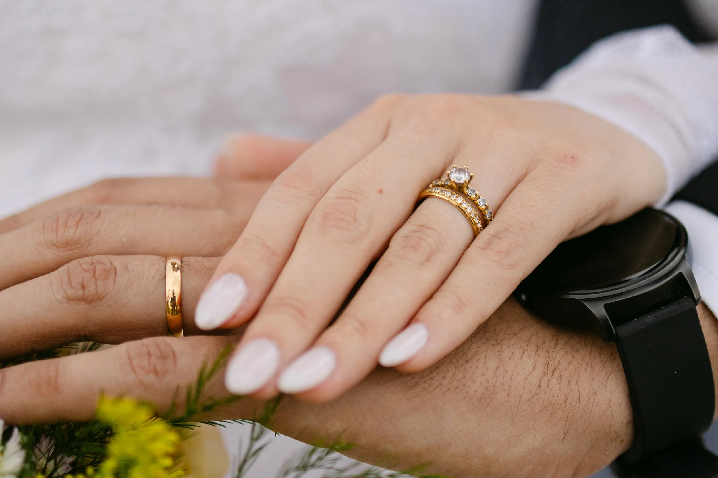 Primer plano de las manos de una pareja entrelazadas, con ambos usando anillos de matrimonio de oro. La mujer tiene uñas pintadas de blanco y lleva un reloj negro en la otra mano. En el fondo, hay flores de colores y un fondo borroso.