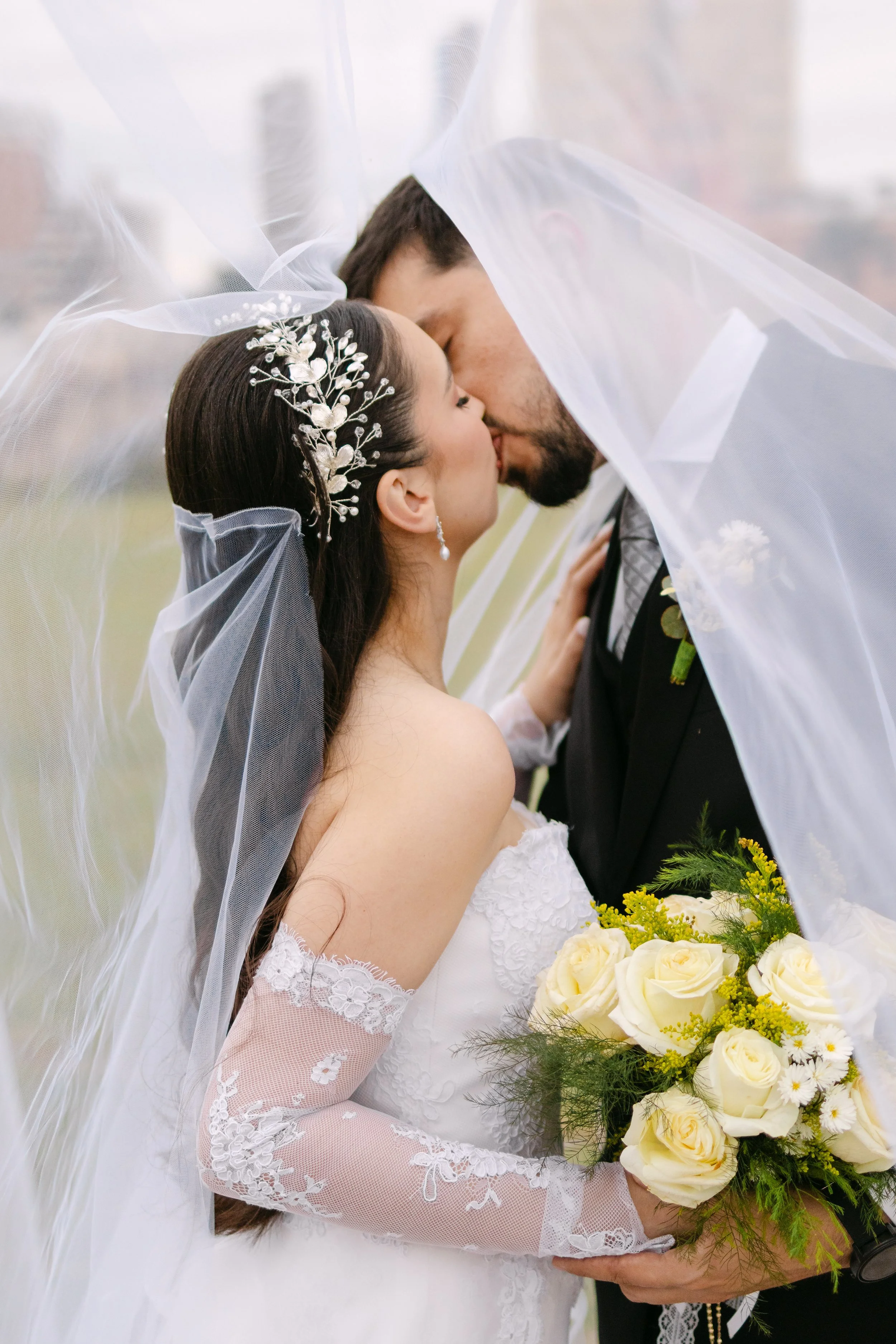 Pareja de recién casados en ceremonia de boda, la novia con vestido de encaje y guantes de encaje, y el novio con traje negro, compartiendo un beso bajo un velo de novia con flores adornando su cabello y sosteniendo ramo de rosas blancas con verdes y