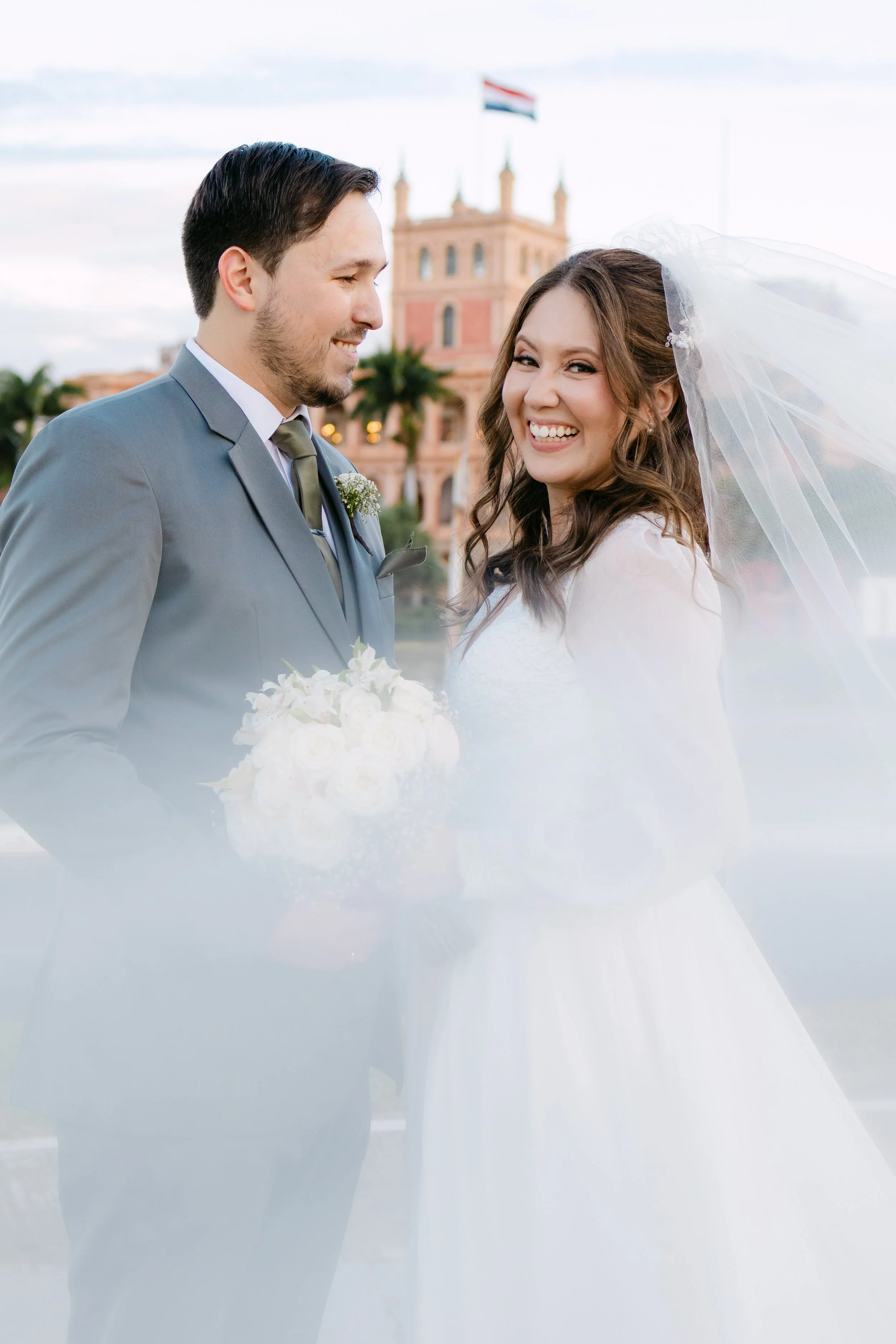 Pareja de recién casados con vestido y traje en un lugar con arquitectura colonial y bandera en el fondo.