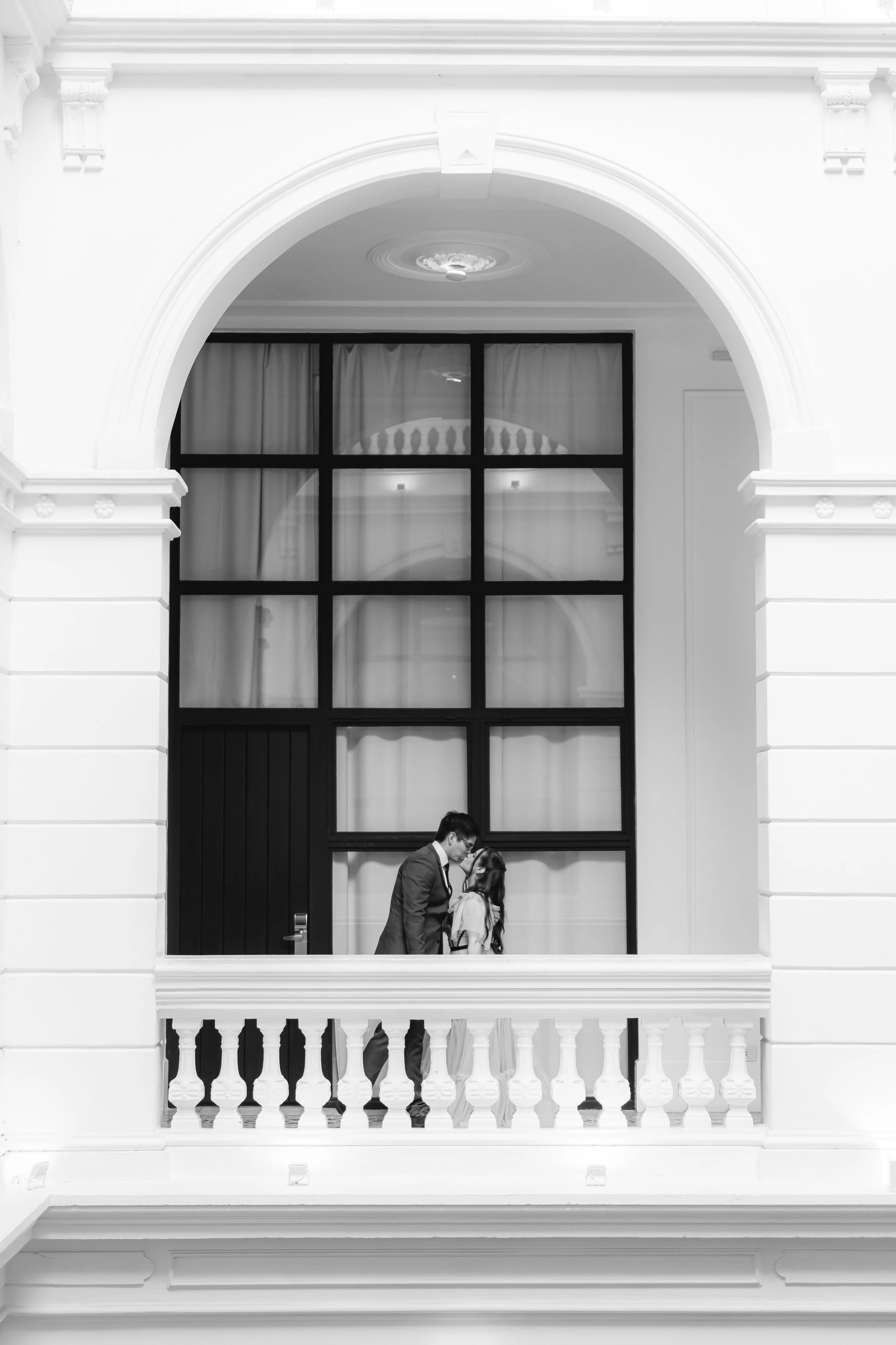 Pareja vestida formalmente, un hombre y una mujer, en el balcón de un edificio histórico, con grandes ventanas y decoración clásica, en un ambiente elegante y romántico.