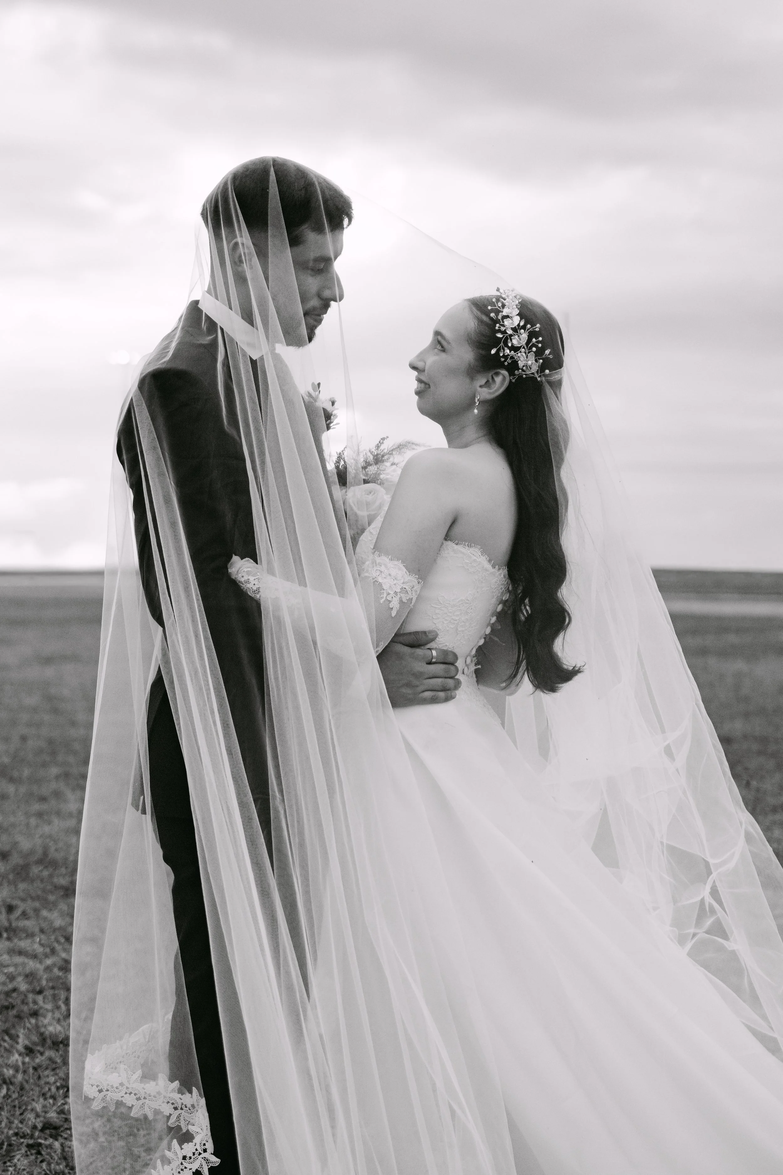 Una pareja de novios en una boda al aire libre, con la novia sonriendo y usando un vestido de novia con encaje y accesorios florales en el cabello, mientras el novio lleva un traje negro, ambos con un velo de novia, en un fondo de cielo nublado.