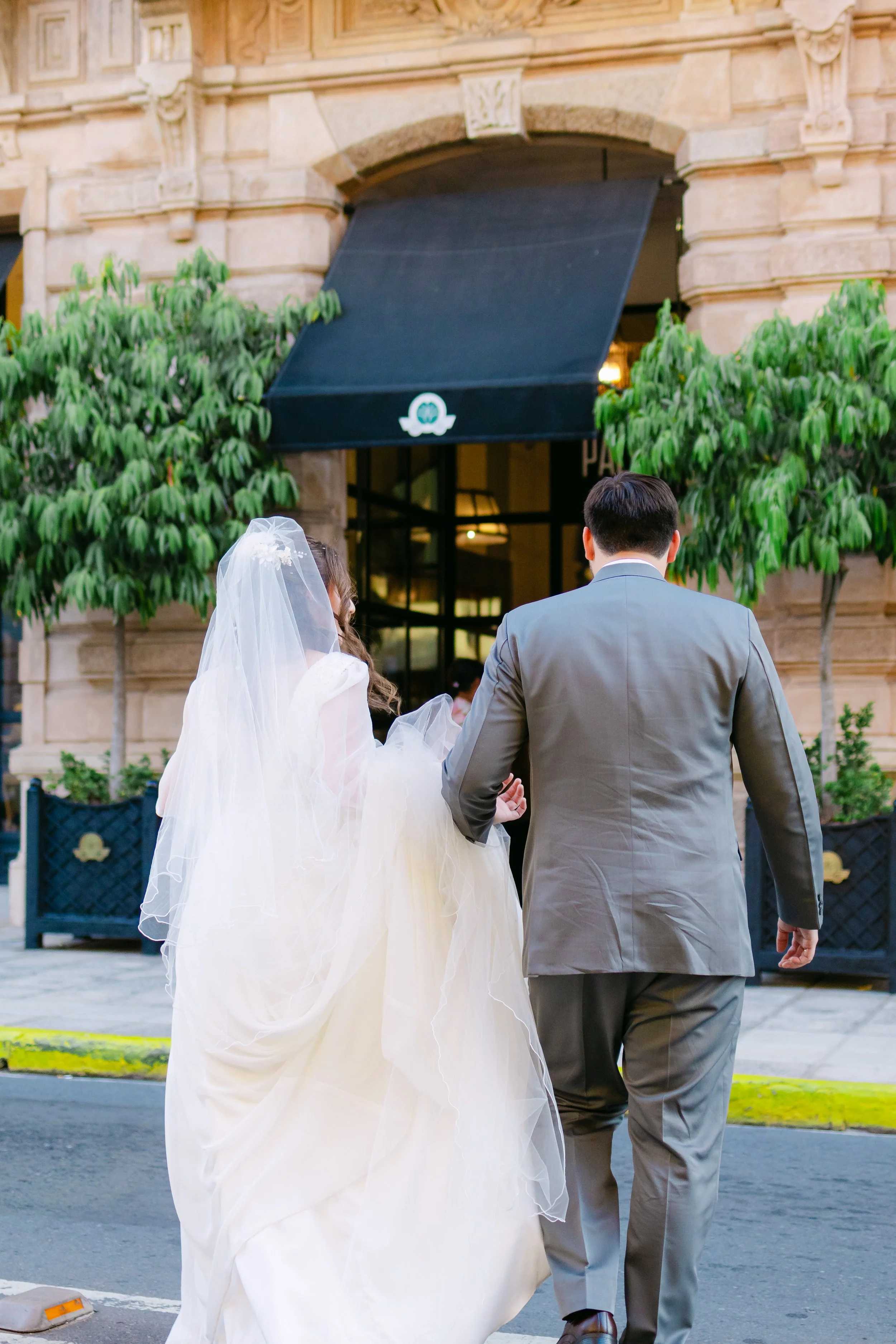 Una pareja de novios caminando por la calle frente a un edificio de piedra, con árboles verdes a ambos lados y una tienda o restaurante con toldo negro en el fondo.