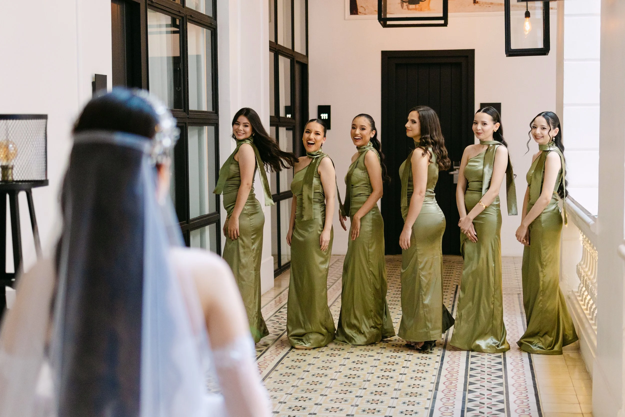 Seis mujeres con vestidos largos de color verde jade posando y sonriendo para una fotografía en un pasillo con pisos de mosaico.