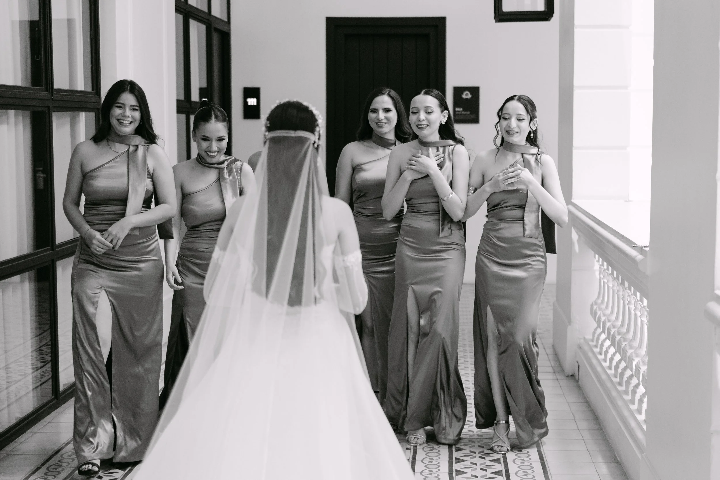 Grupo de cinco mujeres con vestidos elegantes de color oscuro, en una pasarela en un evento, vistas desde atrás, una de ellas con un velo, posiblemente en una boda.
