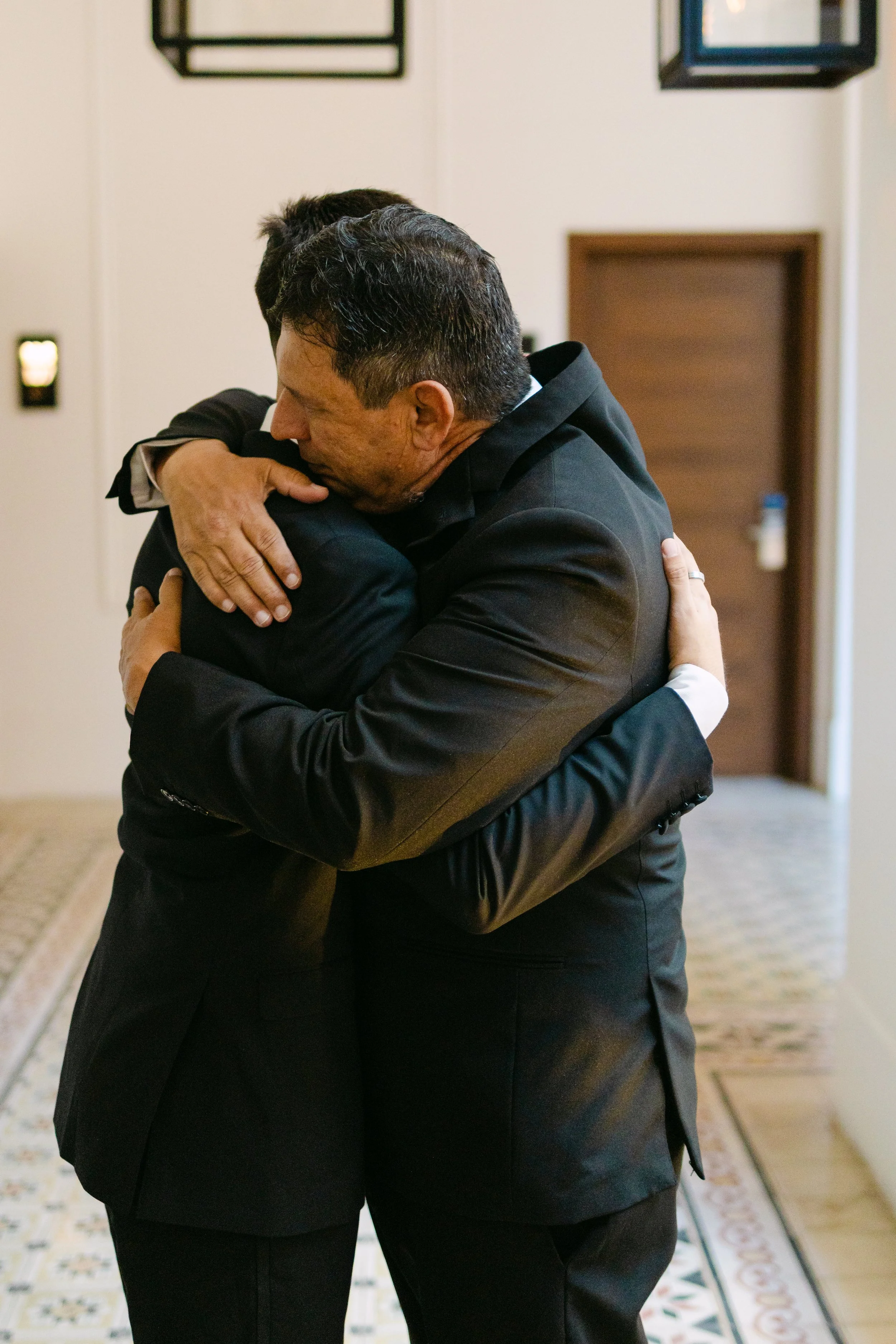 Dos personas abrazándose en un espacio interior con alfombra decorativa y paredes blancas.