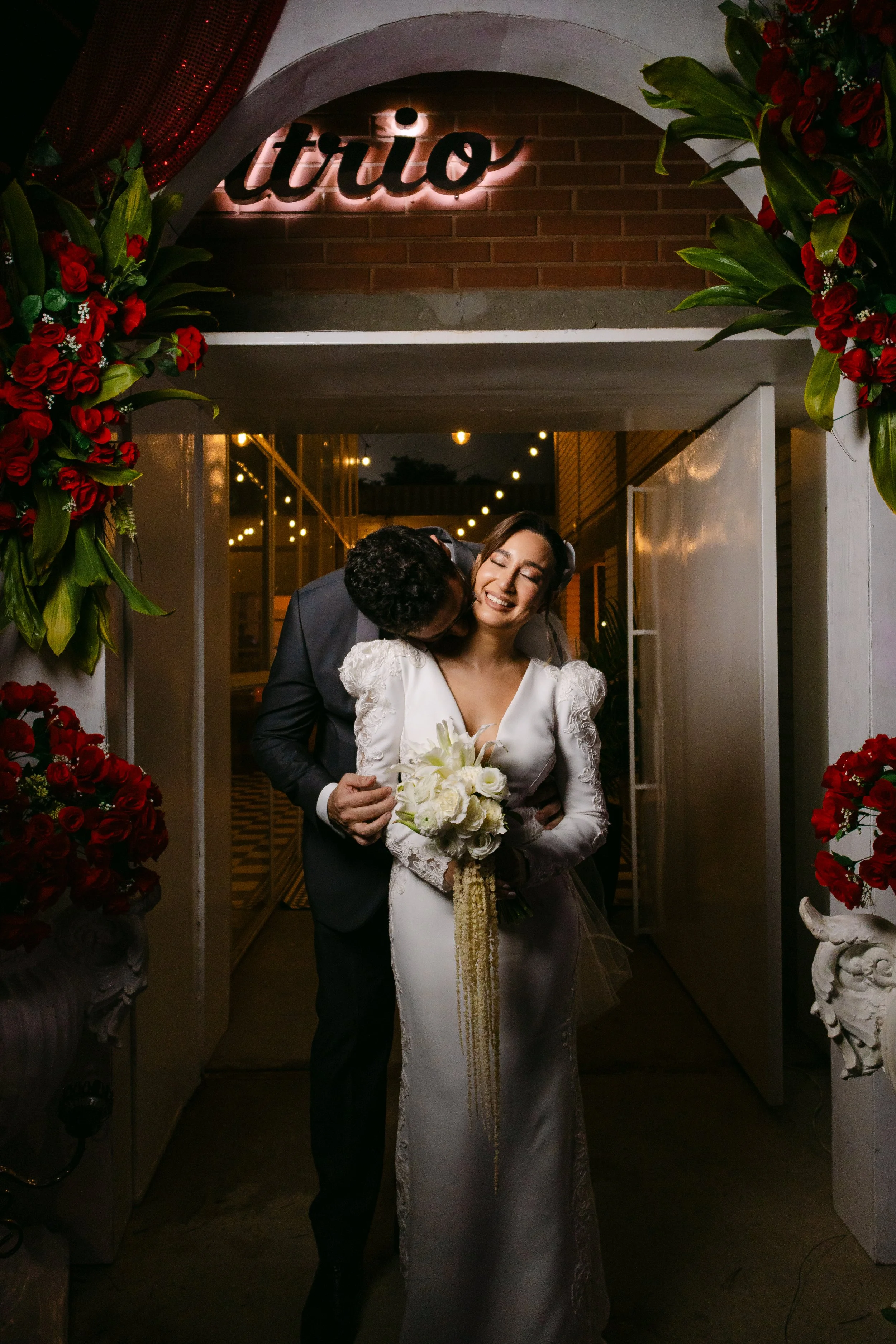 Momento de una boda, una pareja en el umbral de una puerta decorada con flores rojas y verdes, con una señal luminosa que dice 'tuio' en el fondo.