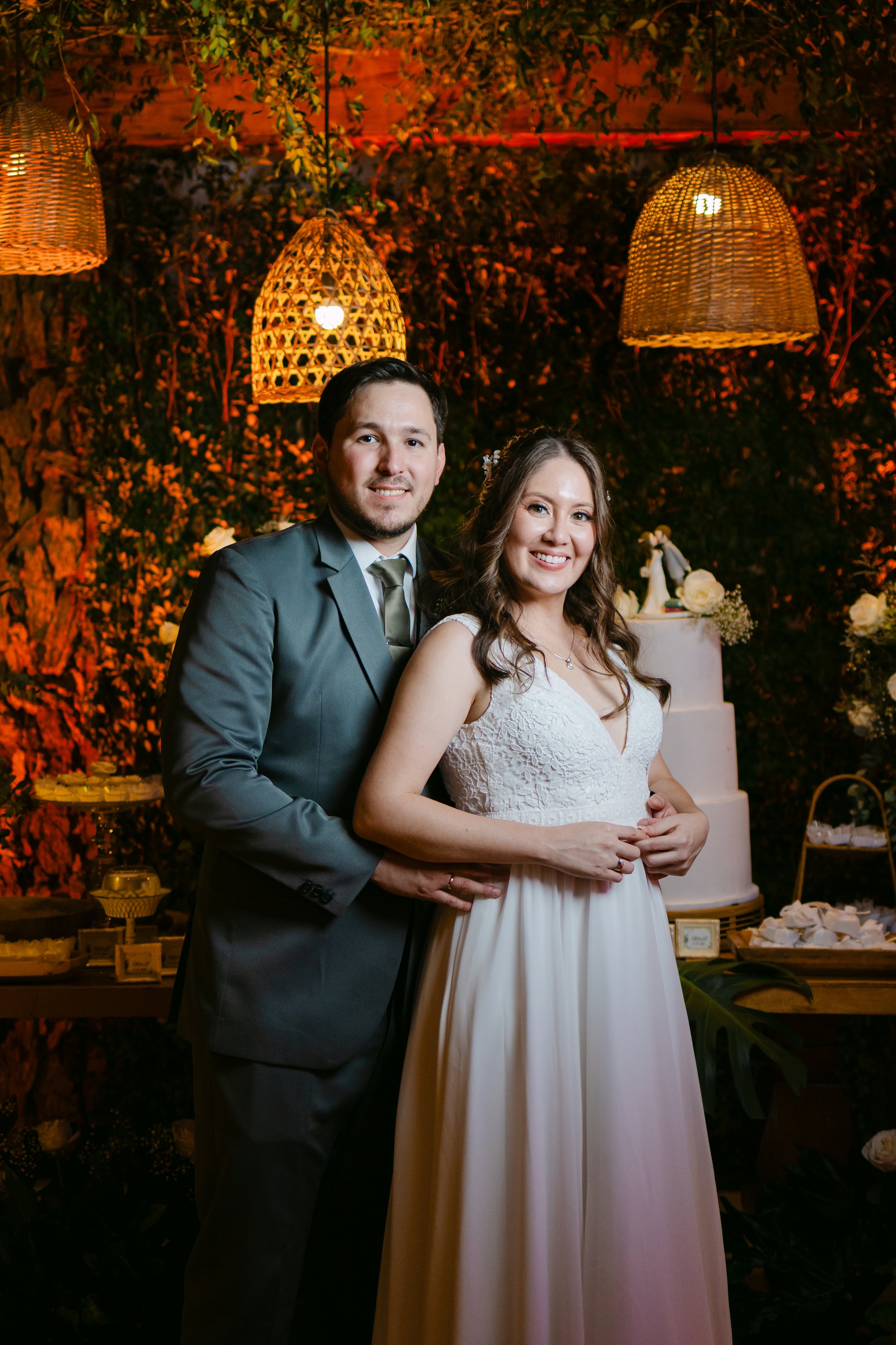 Una pareja de recién casados en su boda, con decoraciones de iluminación cálida y fondo de plantas y flores, frente a un pastel de bodas y mesa decorativa.