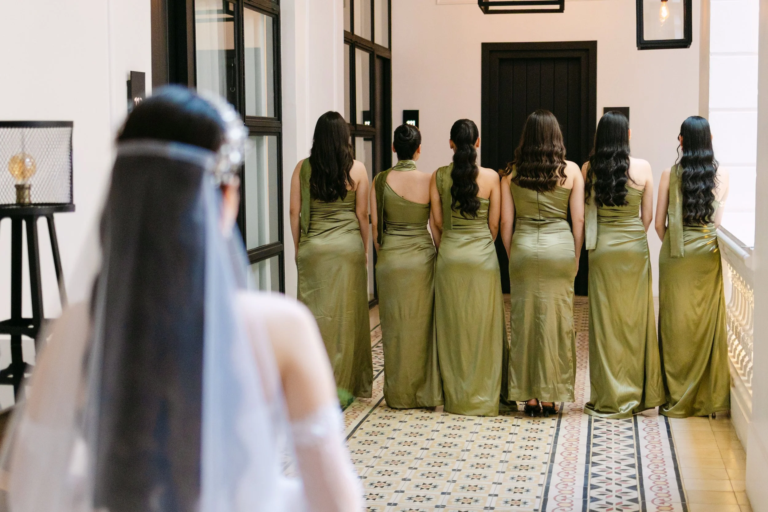 Variedad de mujeres vestidas con trajes largos y de color verde, de espaldas, caminando en línea en un pasillo con piso de azulejos decorativos y paredes blancas, en un espacio interior elegante.