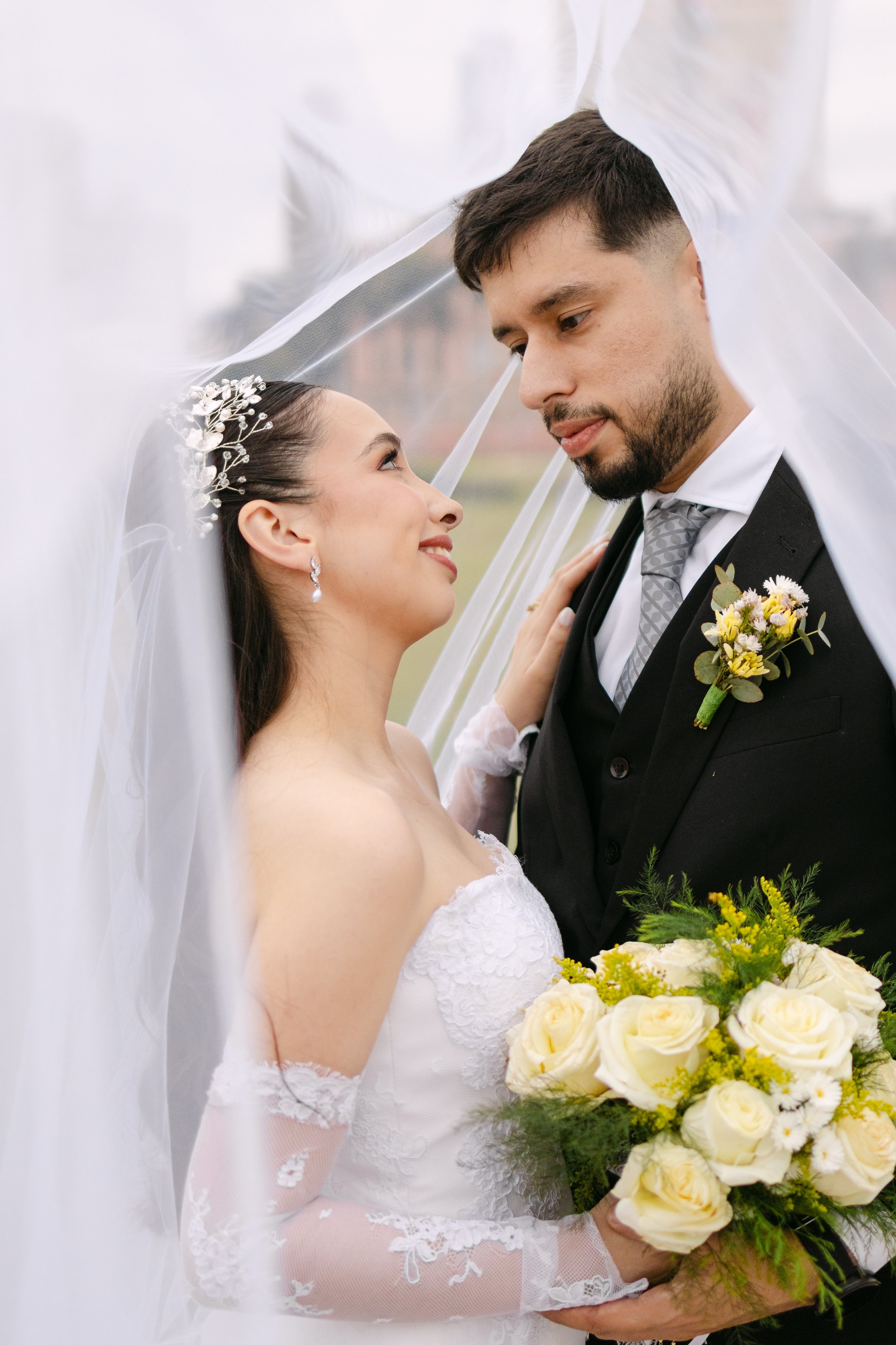 Una pareja de recién casados en una boda, la novia vestida con vestido blanco y el novio con traje negro y corbata, sosteniendo un ramo de flores blancas y amarillas.