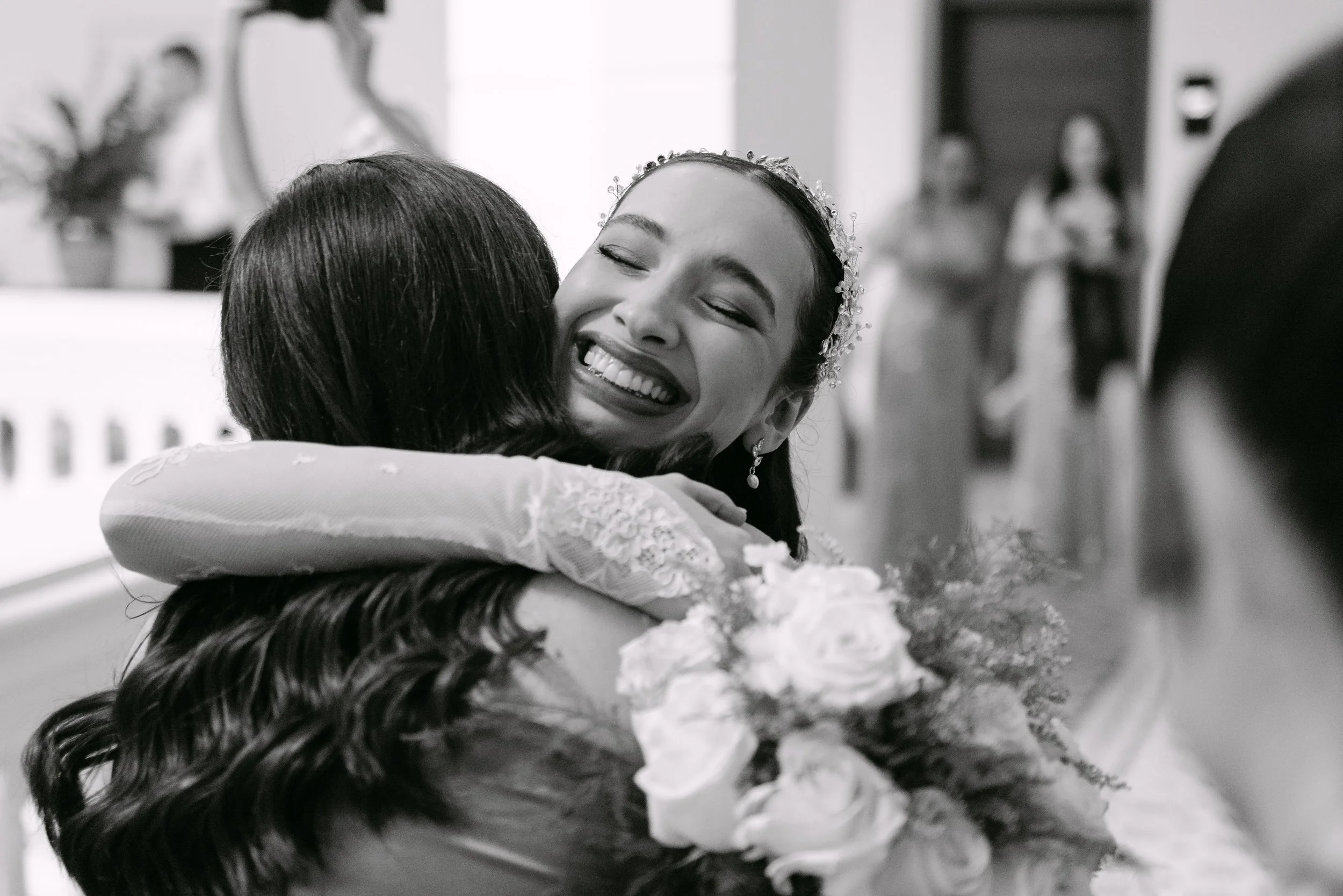 Mujer sonriendo y abrazando a otra persona en una celebración, con flores en el fondo.