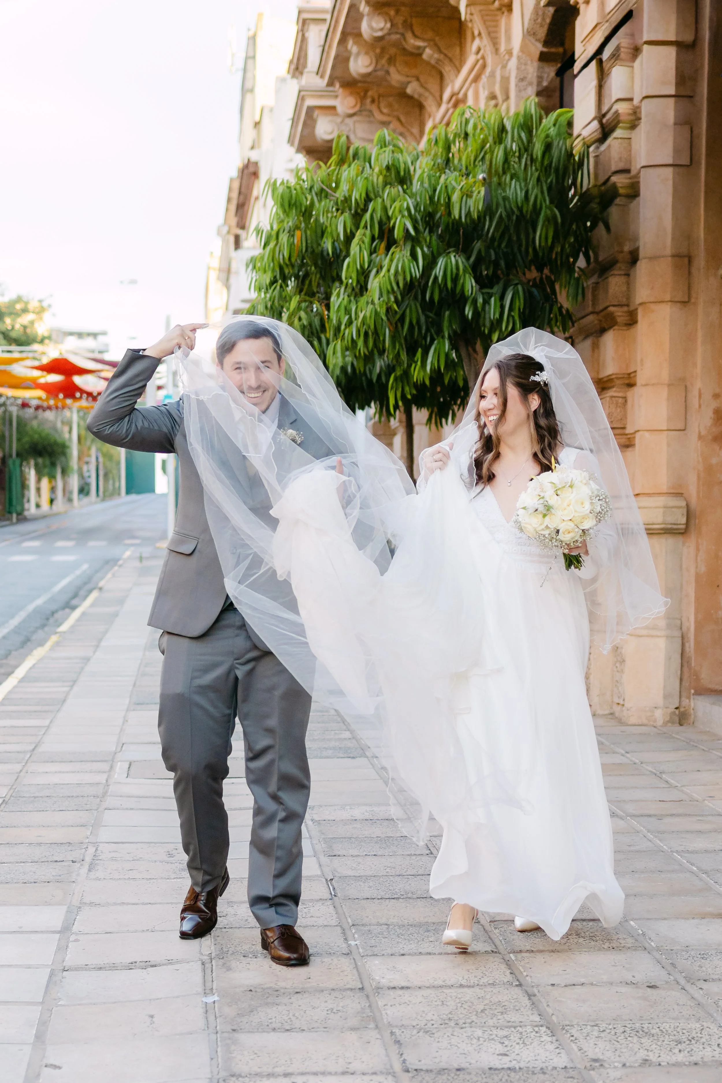Una pareja de recién casados caminando por la calle, ambos con velo de novia y la novia sosteniendo un ramo de flores blancas.