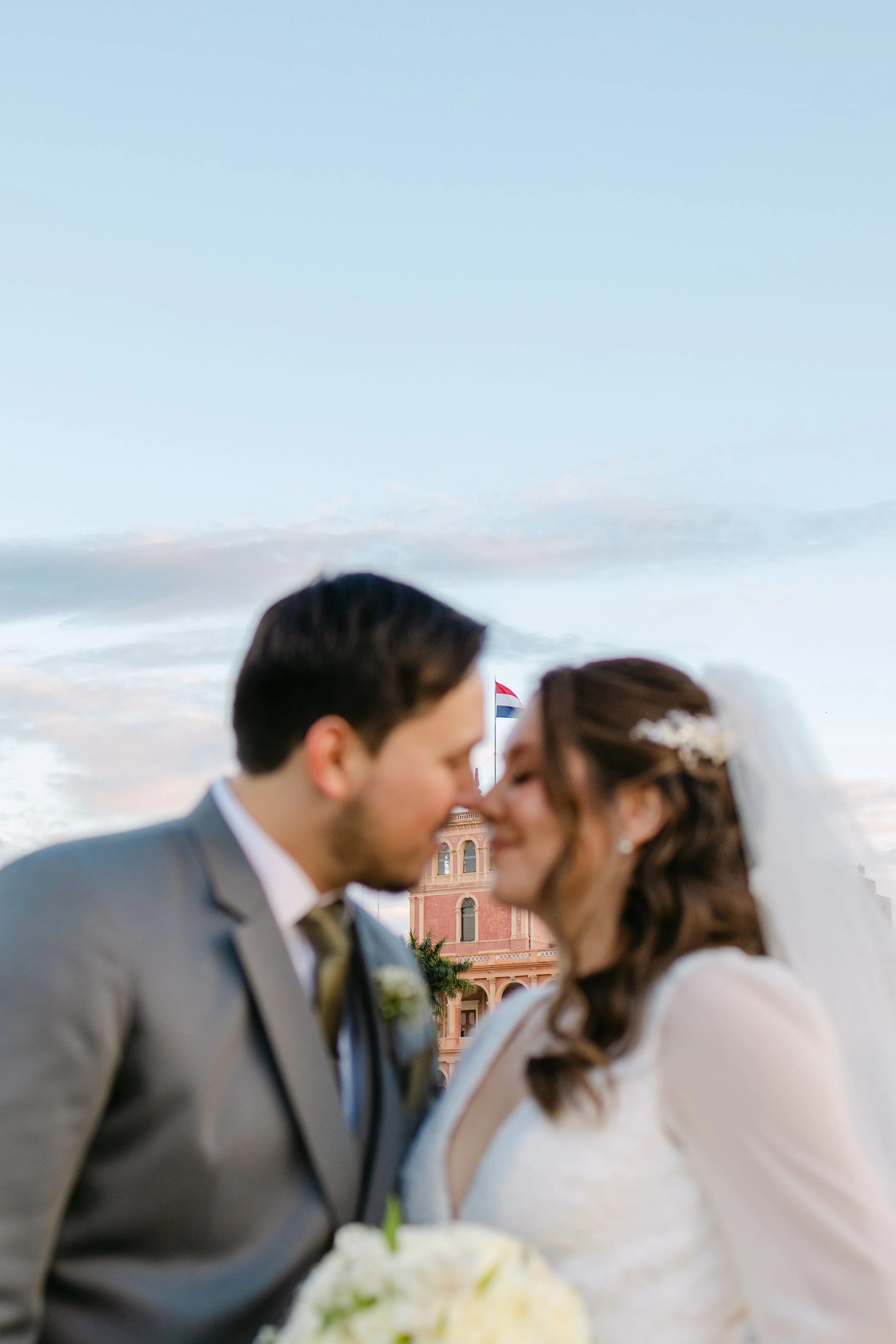 Pareja de recién casados en un beso, con un edificio histórico y una bandera en el fondo, durante el atardecer.