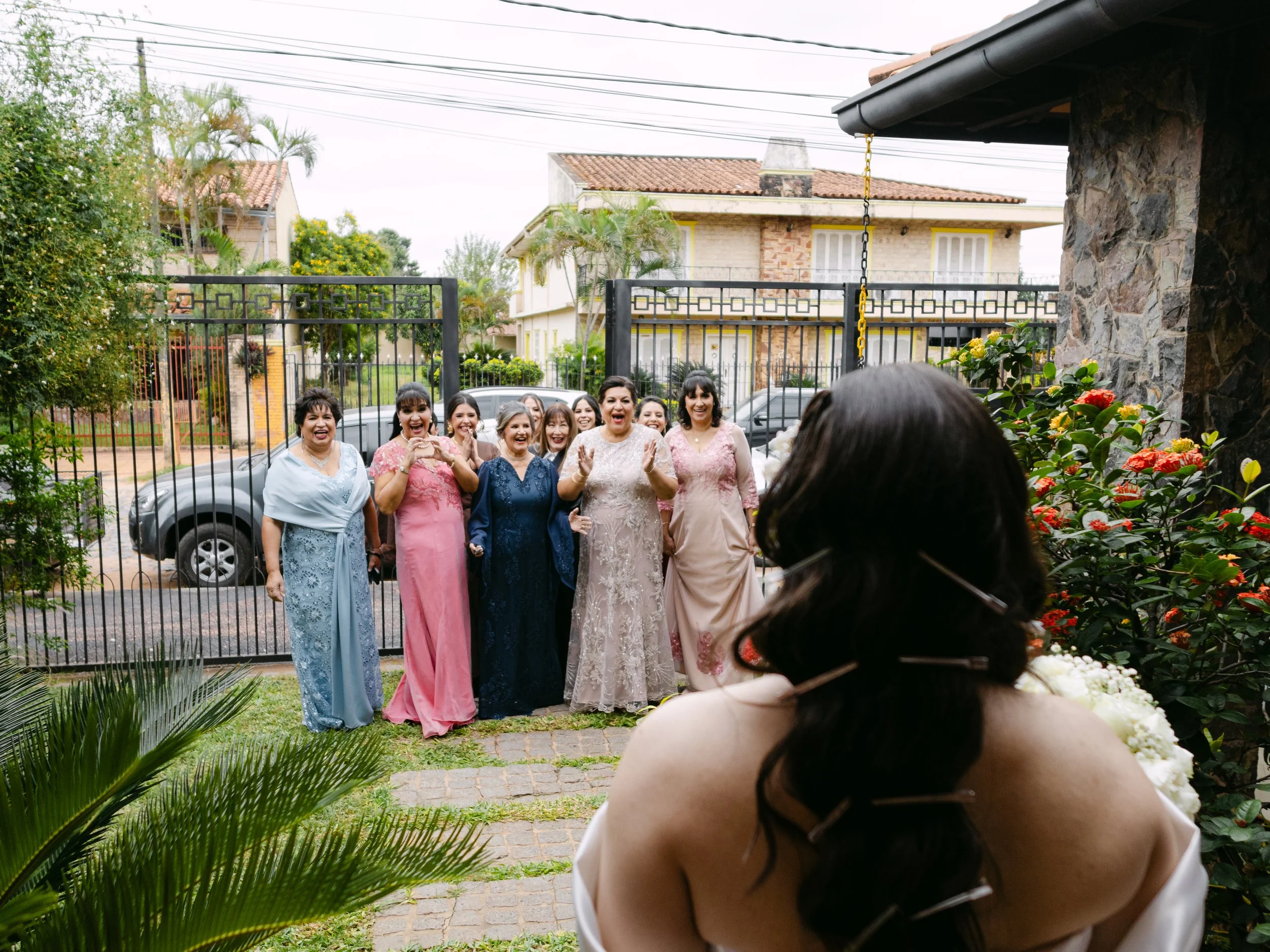 Grupo de mujeres en ropa elegante sonriendo en un evento al aire libre en una entrada de casa rodeada de plantas y flores.