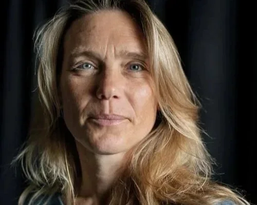 Portræt af en kvinde med langt, blond hår, blå øjne og en let smuksminke, mod en mørk baggrund.