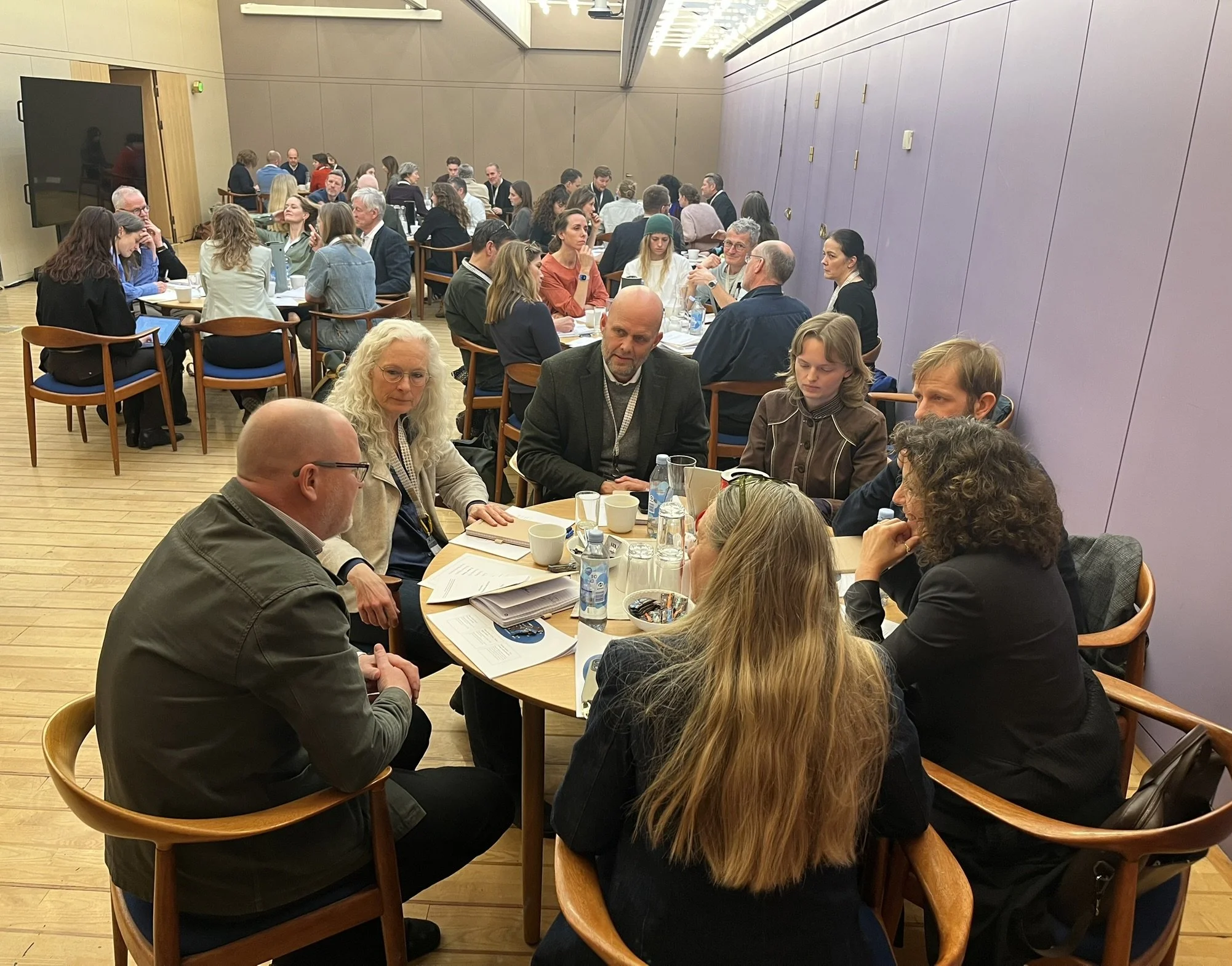 Rundbordssamtaler ved Symposium i Forum for En Ny Grøn Oplysningstid. En stor gruppe mennesker sidder ved flere borde i et konferencerum, nogle taler, andre læser eller bruger deres telefoner, med papir, kopper og drikkevarer på bordene.