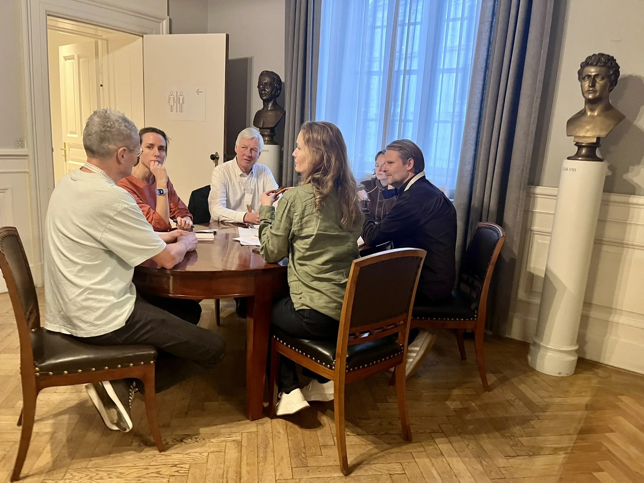 Rundbordssamtaler ved Symposium i Forum for En Ny Grøn Oplysningstid. En gruppe mennesker i samtale rundt et rundt bord i et indendørs rum med store vinduer og to skulpturer.