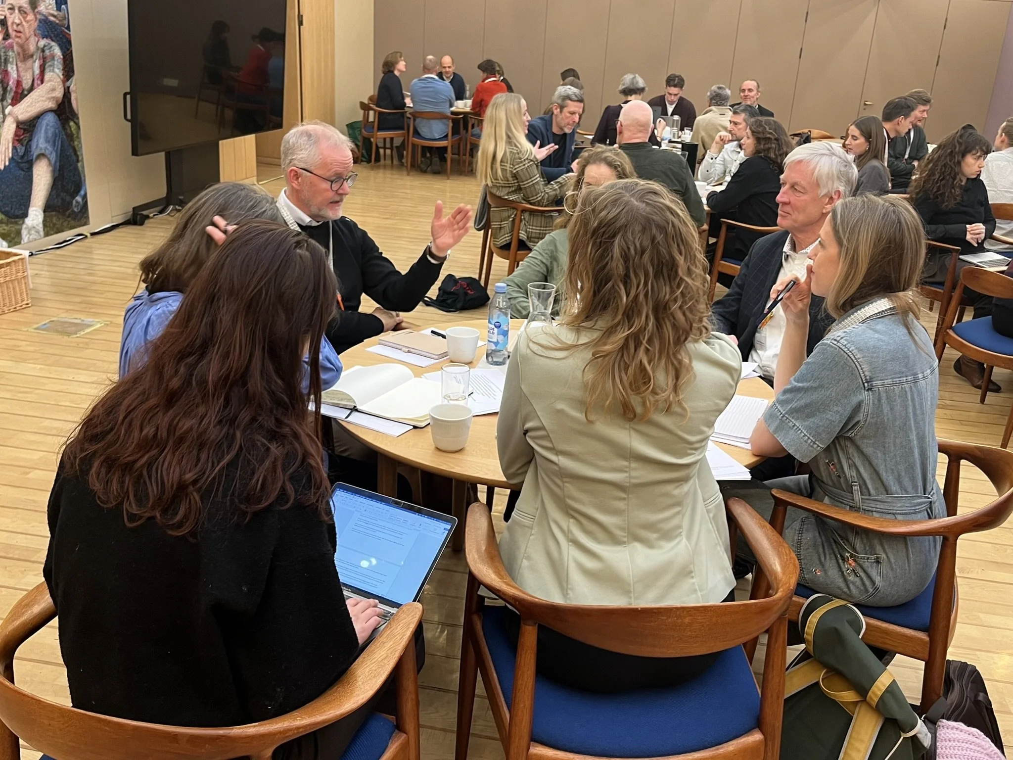 En gruppe mennesker sidder omkring et rundt bord i en indendørs mødesal, nogle er i samtale, og én person bruger en laptop. Alle er fokuserede, og der er workshops- eller mødematerialer på bordet.