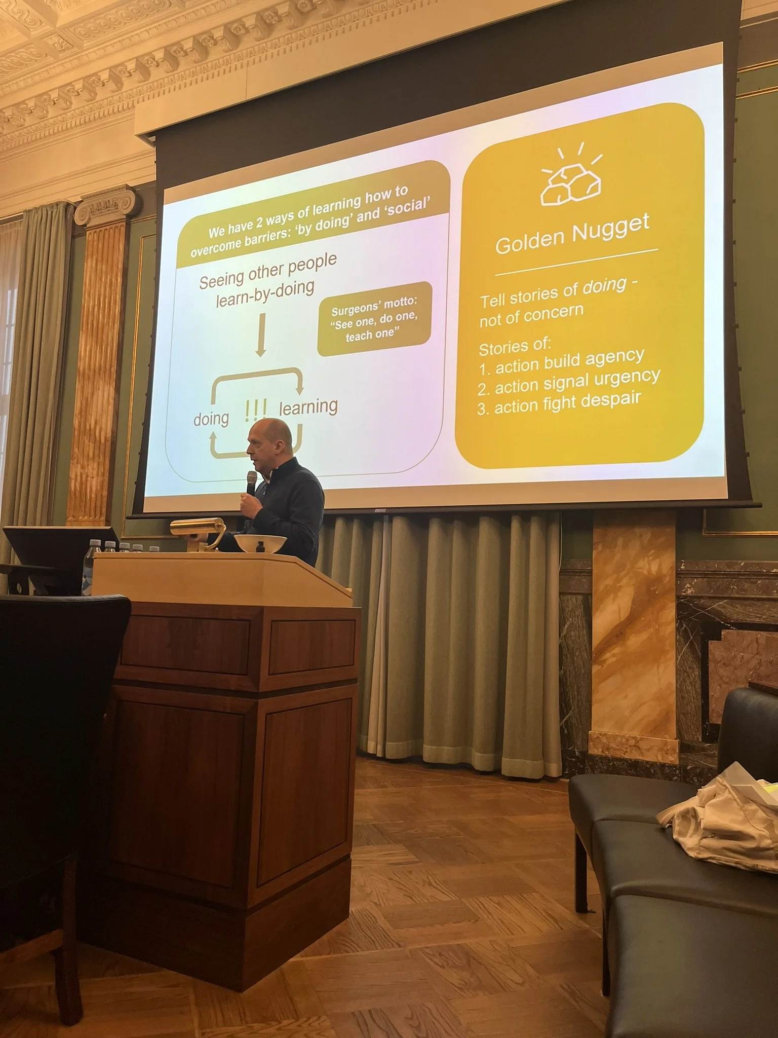 Et foredrag i et elegante lokale. Der er en stor projektor, der viser en præsentationsside med baggrundsfarve i hvid, og teksten handler om at overvinde barrierer gennem 'doing' og 'social'. Der står også en gule boks med 'Golden Nugget' og tips til at fortælle historier om handling, ikke bekymring, og tre typer historier: handling, signal, kamp.