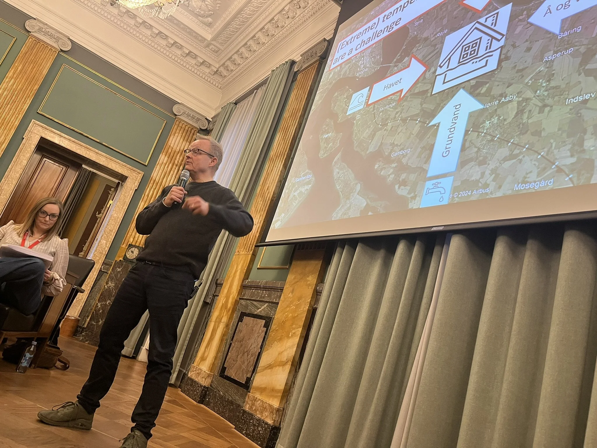 Et gut ved et foredrag med en forsker i havvandsstigninger, professor Sebastian Mernild,  i en elegant, klassisk salon med grønne vægge og gulddetaljer. En mand holder mikrofon og taler, mens en kvinde sidder i forgrunden med papirblok.