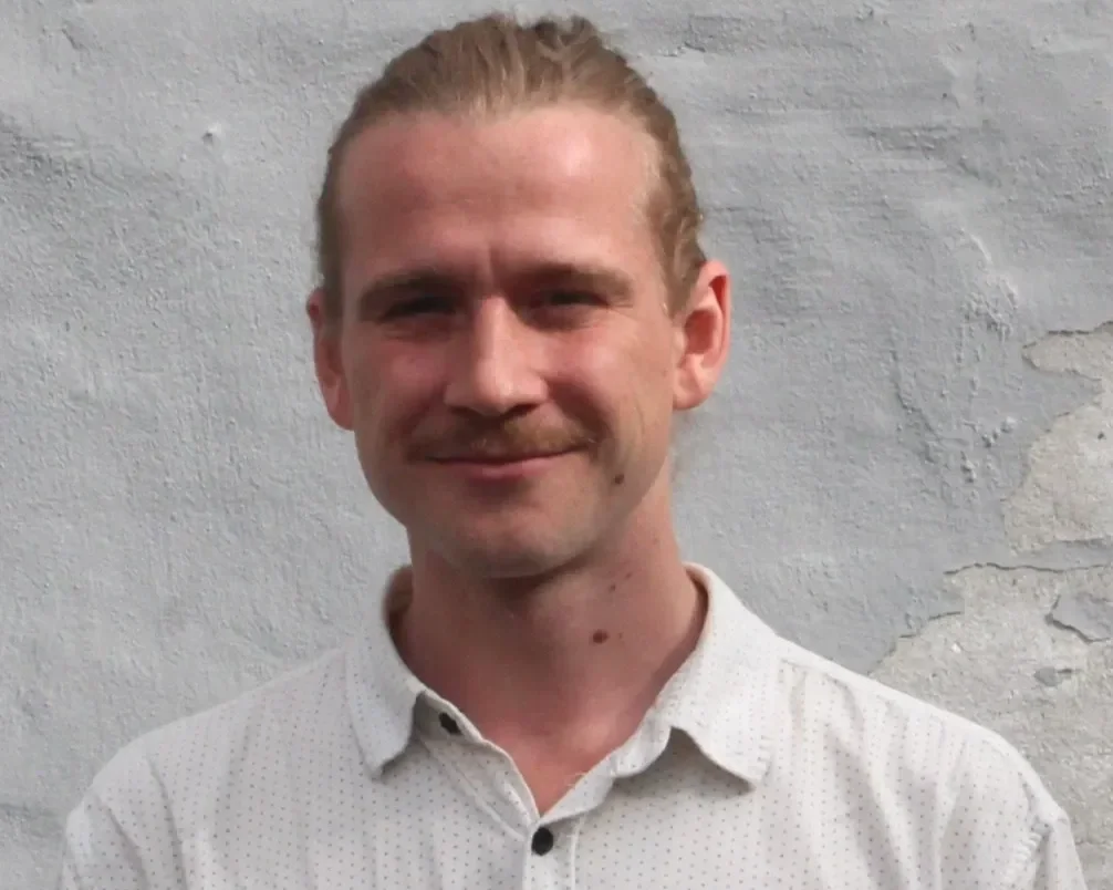 En mand med lyst hår, skæg og blå øjne, der smiler, iført en hvid skjorte, står foran en grå væg.