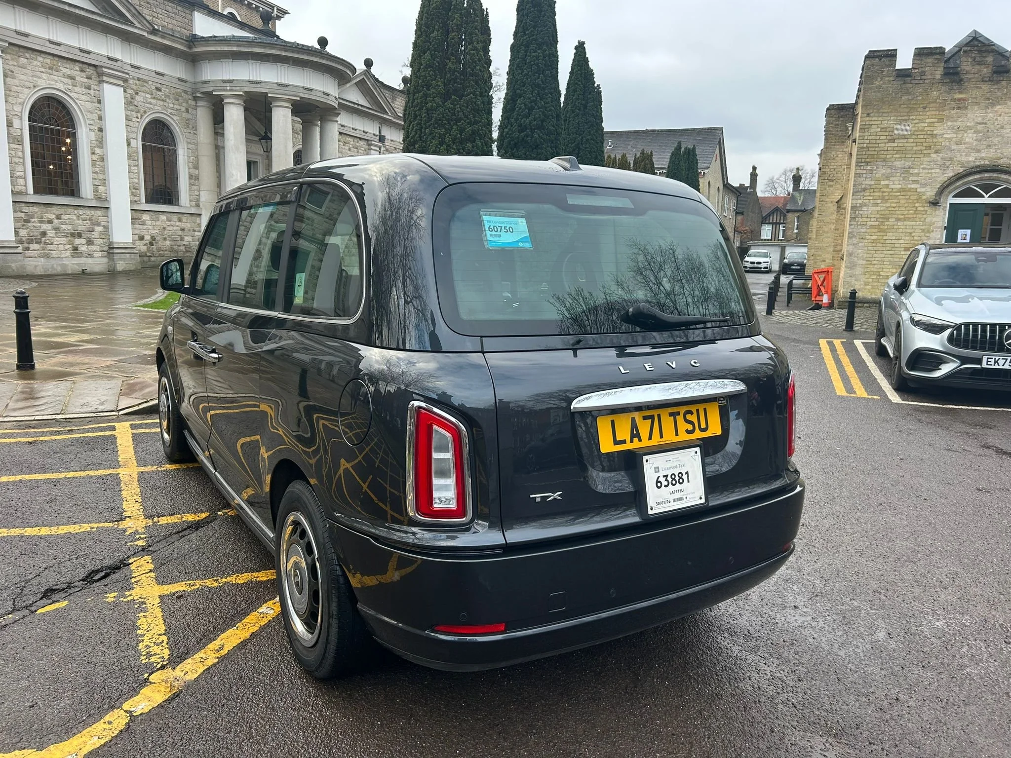 Black Leyc taxi with the license plate LA71 TSU parked in a designated parking space next to a grey car, in front of historic stone buildings with arched windows and tall trees, on a wet pavement on a cloudy day.