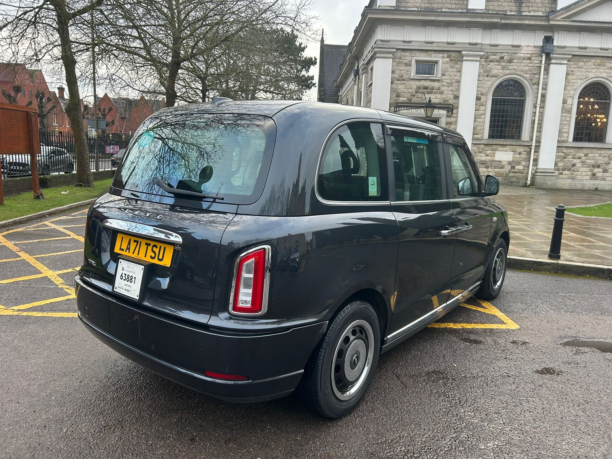 A black London taxi cab parked in a designated parking spot on a wet pavement, with a historic stone building and leafless trees in the background.