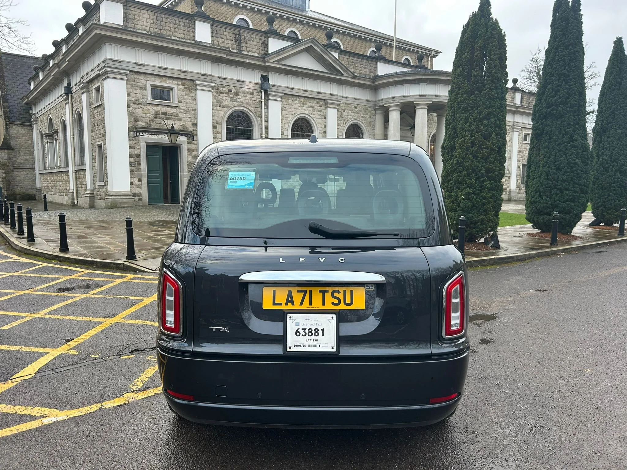 Back view of a black taxi cab with a UK license plate parked on the street in front of an elegant stone building with columns and a group of tall, dark green trees.