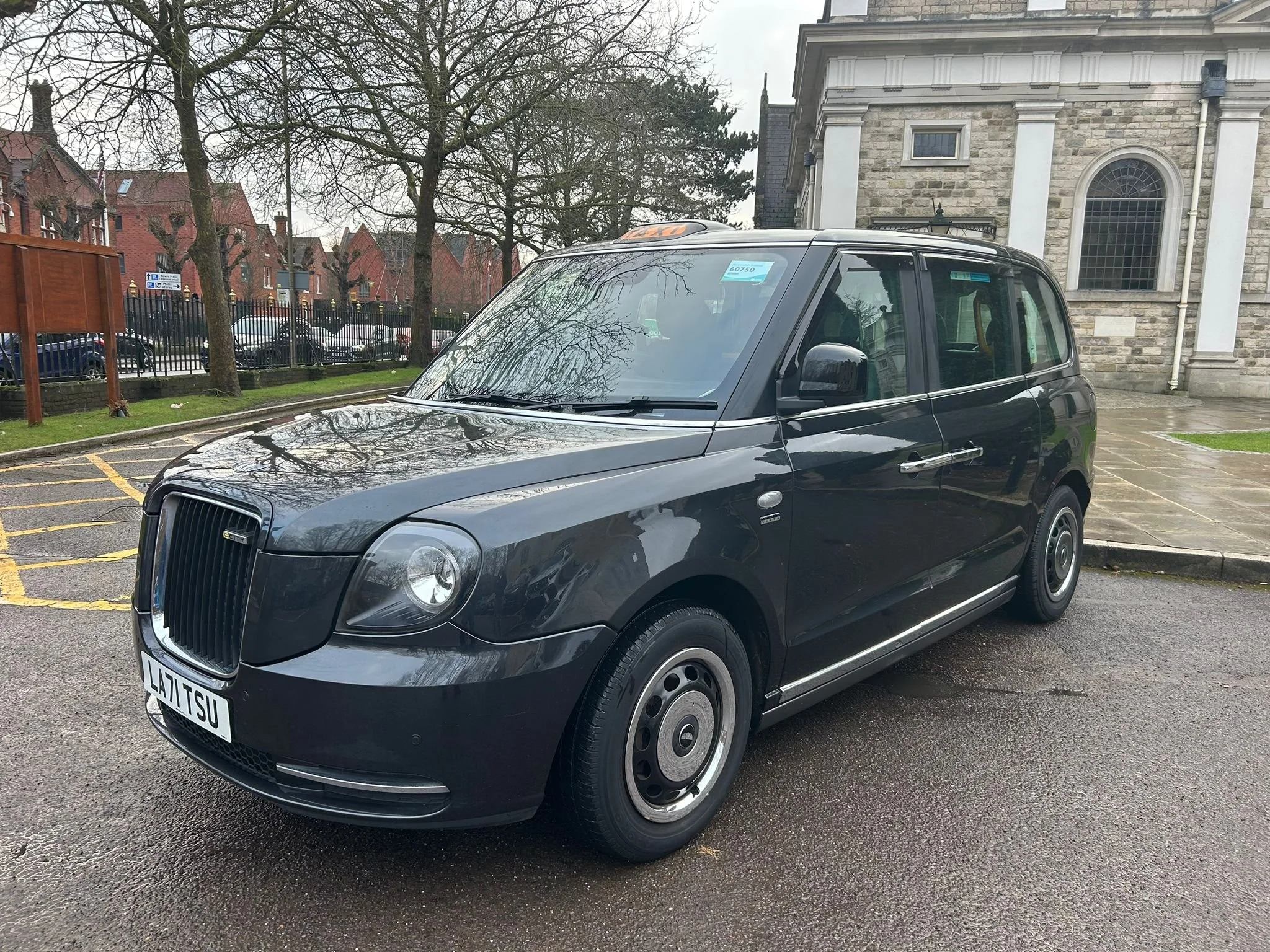 Black London taxi parked in front of a stone building with large arched window, leafless trees, and other parked cars in the background on a cloudy day.