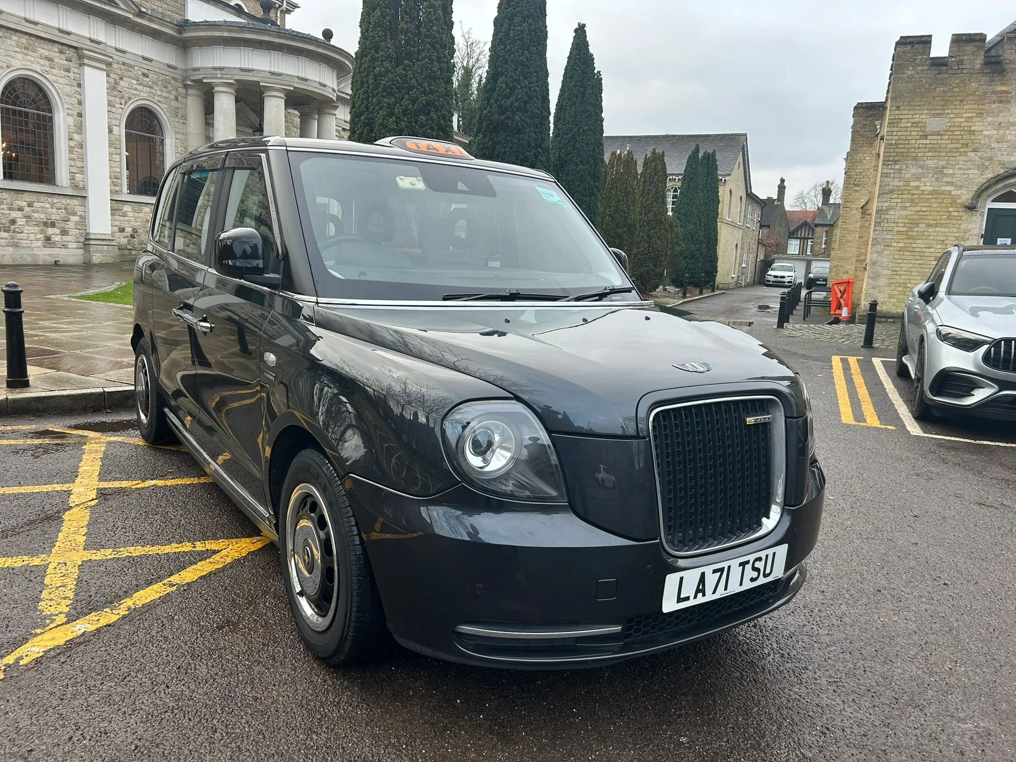 Black taxi cab parked in a designated spot on a wet street in front of historical-style buildings with arched windows, trees, and other parked cars nearby.