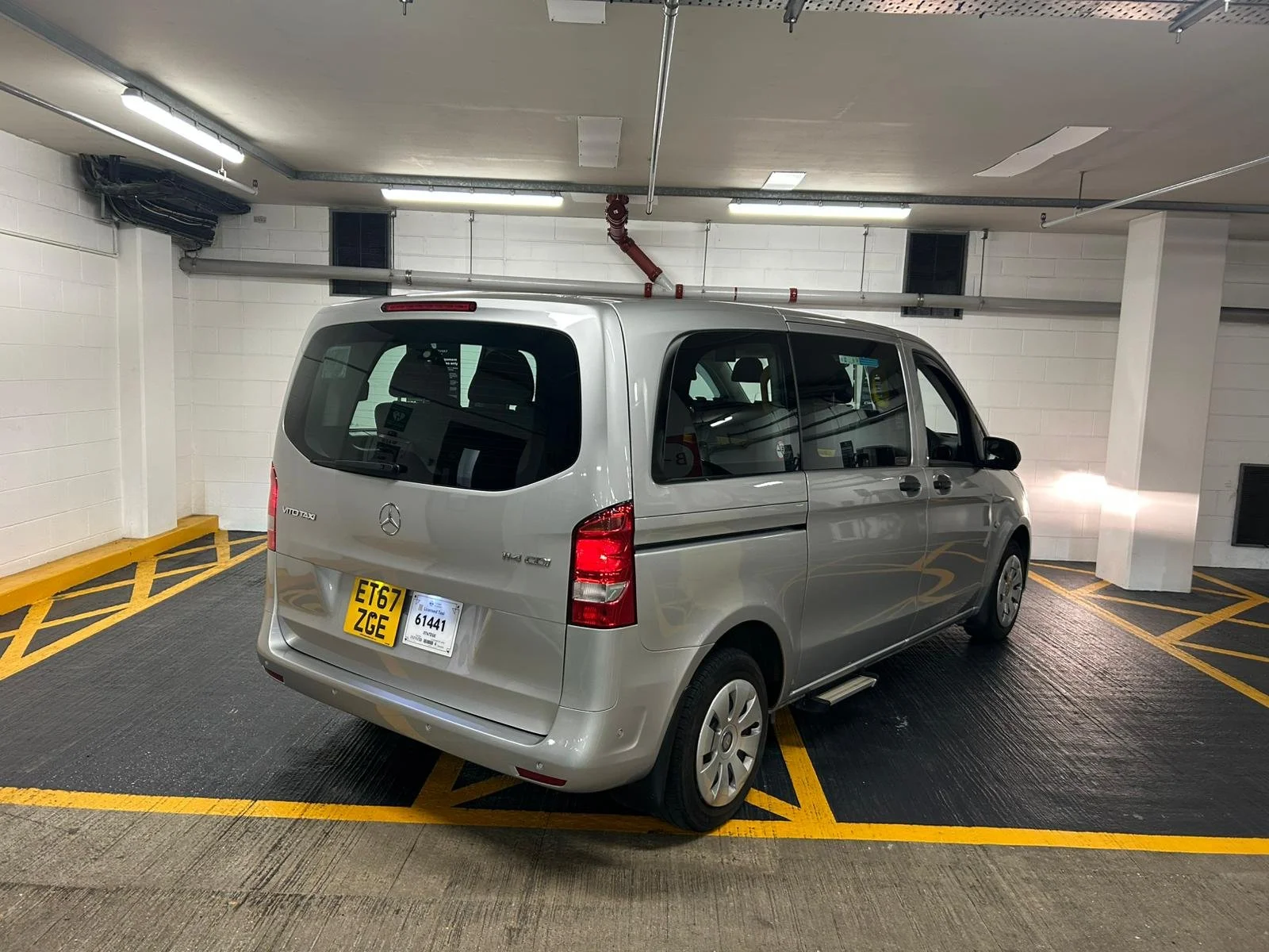 A silver Mercedes-Benz Vito Taxi minivan parked in a designated parking spot in an indoor parking garage with white walls and overhead fluorescent lighting.
