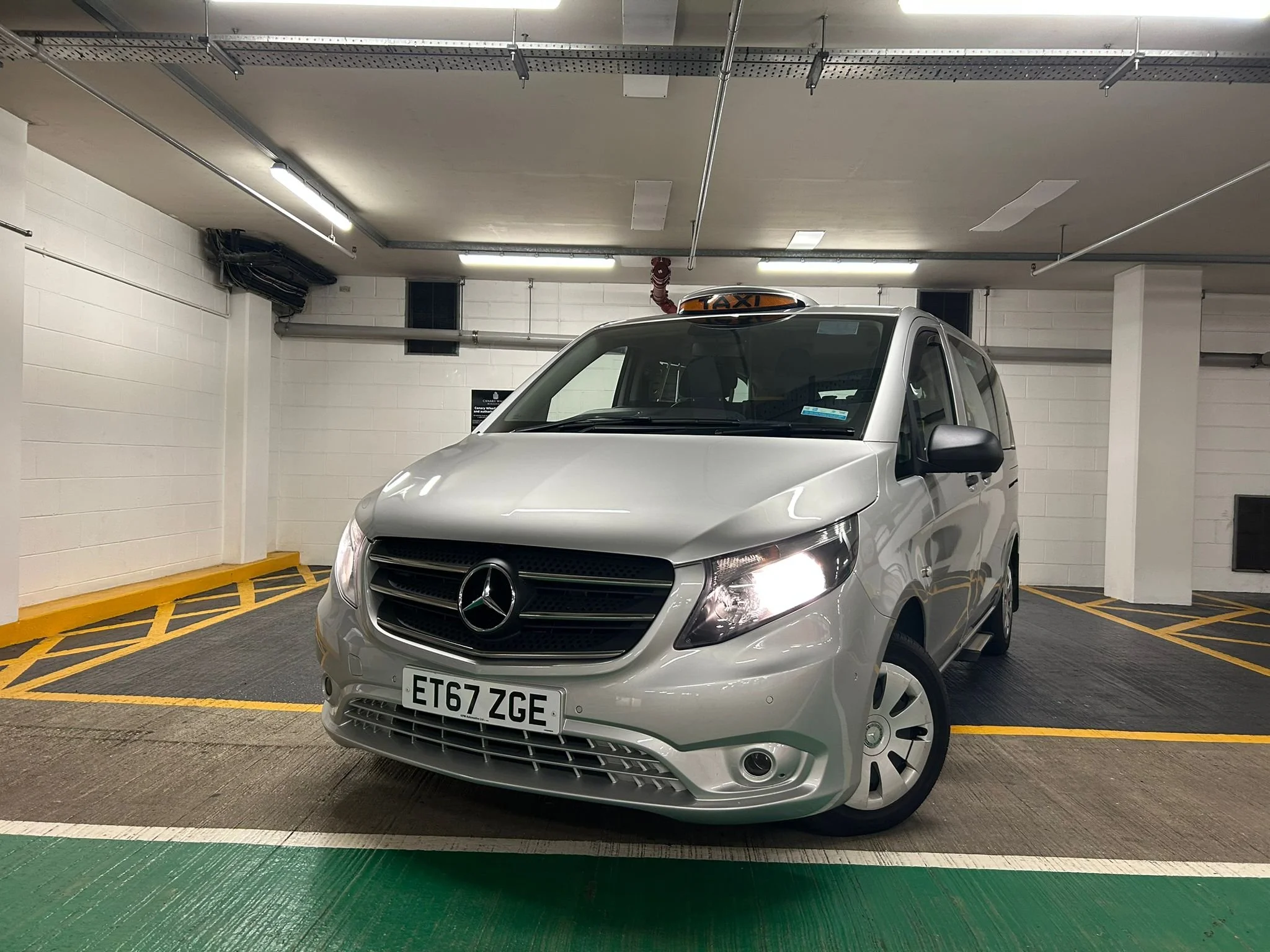 Silver Mercedes-Benz taxi van parked indoors in a parking garage with white walls and fluorescent lighting, front view