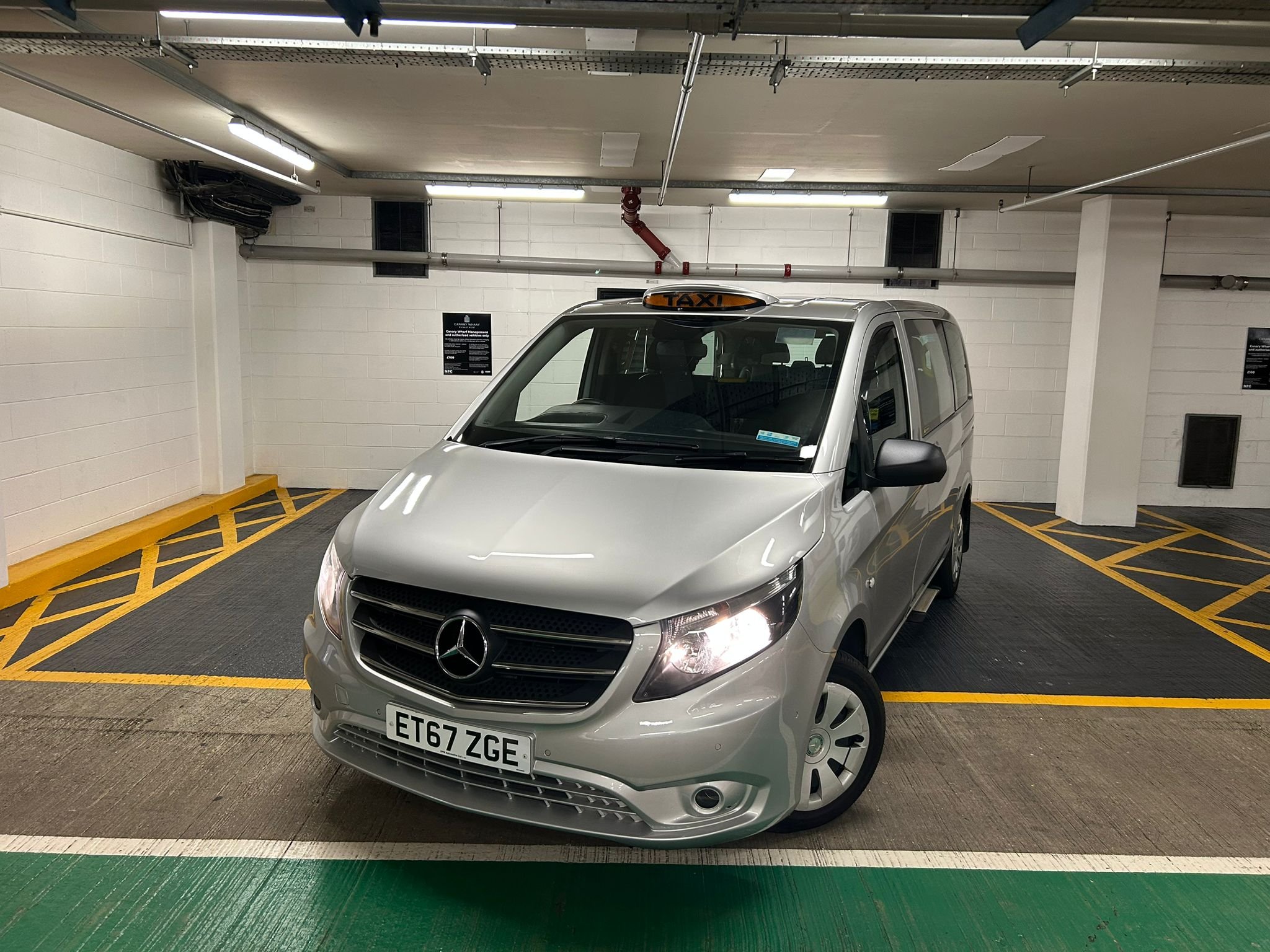 Silver Mercedes-Benz taxi van with lit yellow 'TAXI' sign on top, parked in an underground parking garage.