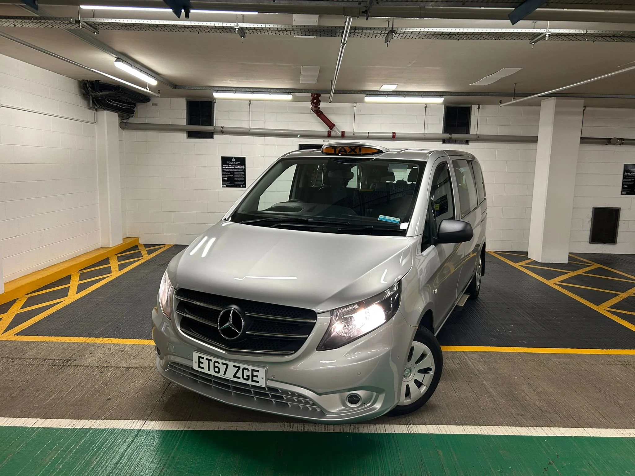 Silver Mercedes-Benz taxi van parked in an indoor parking garage with a 'TAXI' sign on top.