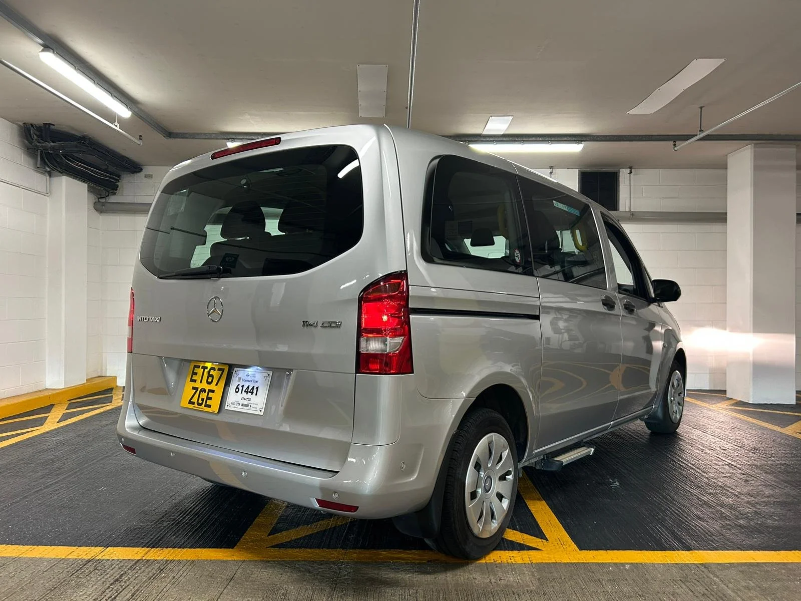 Silver Mercedes-Benz Vito Taxi van parked in an indoor parking garage with yellow parking lines and white painted brick walls.