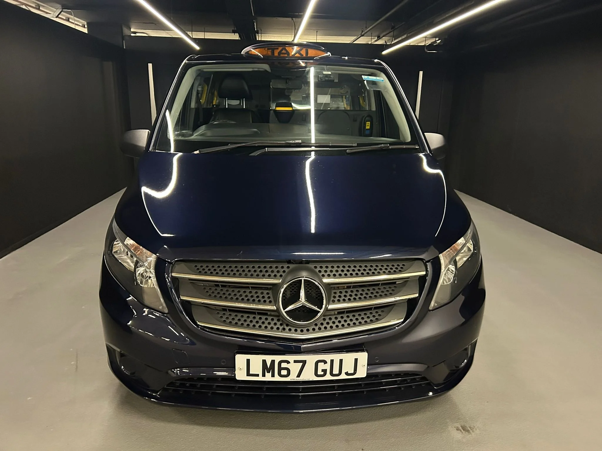 Front view of a dark blue Mercedes-Benz taxi parked indoors, with a visible taxi sign on the roof and a UK license plate reading 'LM67 GUJ'.