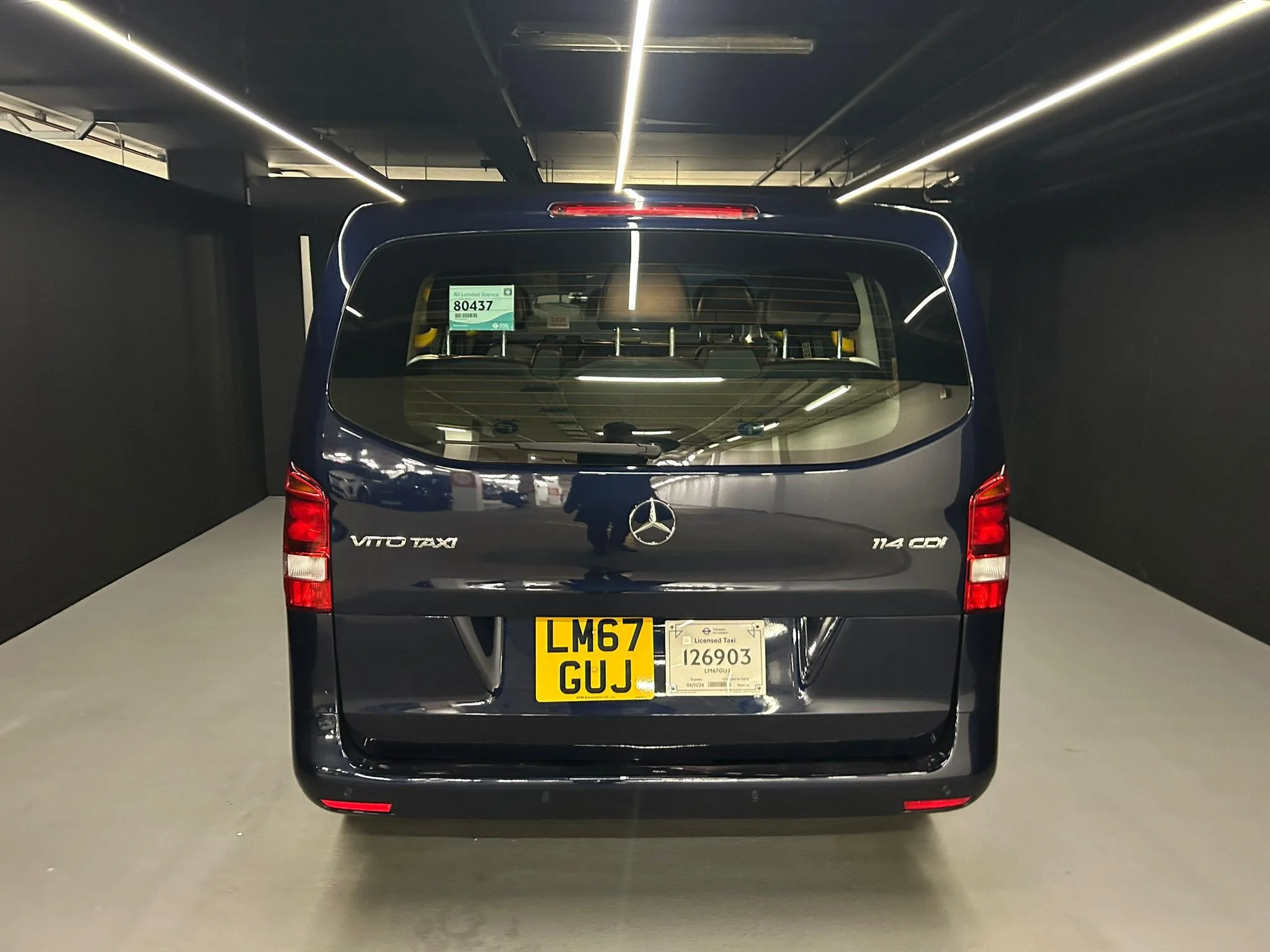 Black Mercedes-Benz Vito Taxi van parked inside a well-lit indoor parking lot with black walls and ceiling.