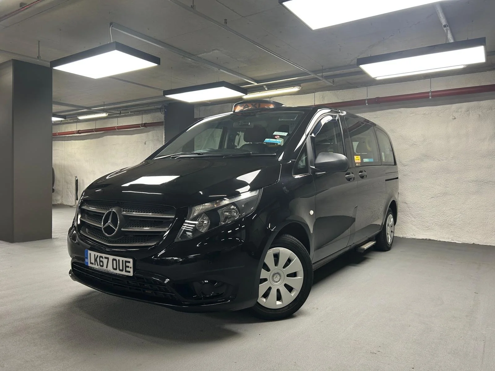 Black Mercedes-Benz van parked in an underground parking garage with white walls and fluorescent ceiling lights.