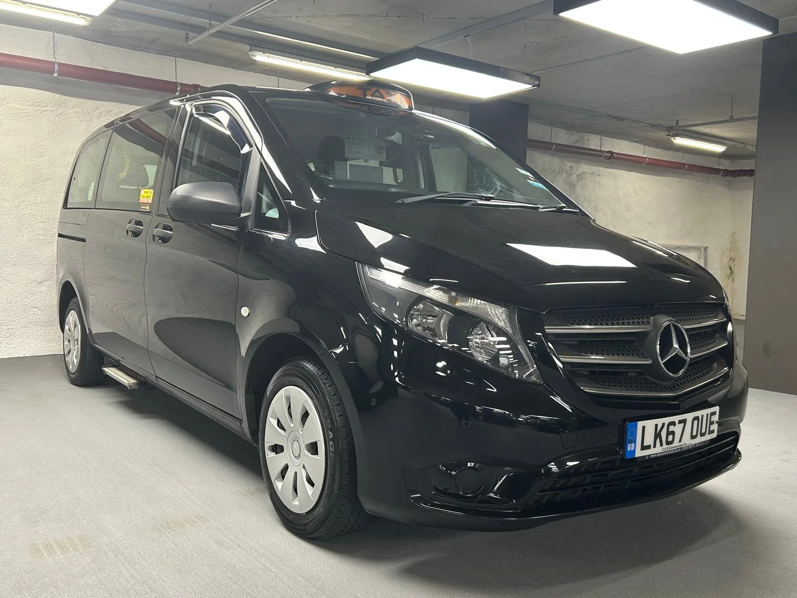 A black Mercedes-Benz van with a taxi light on the roof, parked in an indoor parking garage.
