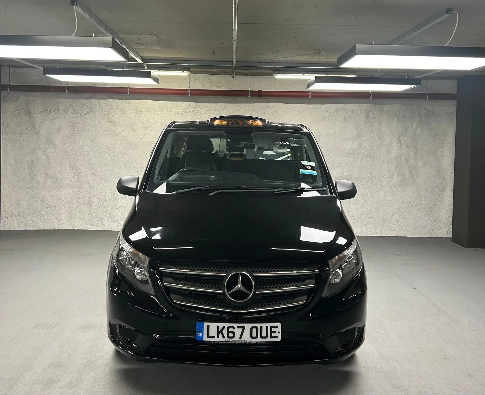 Black Mercedes-Benz taxi van parked in an indoor parking garage under ceiling lights.