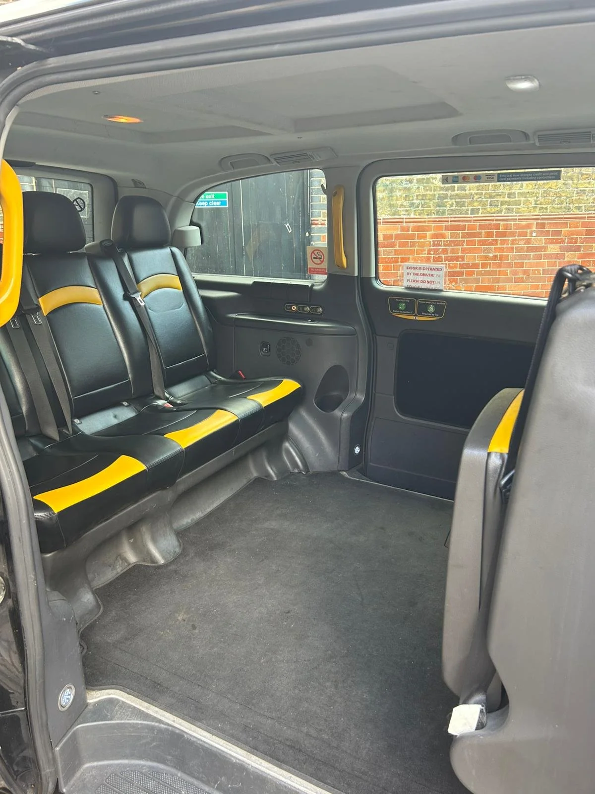 Interior of a passenger van with three black seats with yellow accents on the left side, a black floor, and a window showing a brick wall outside.