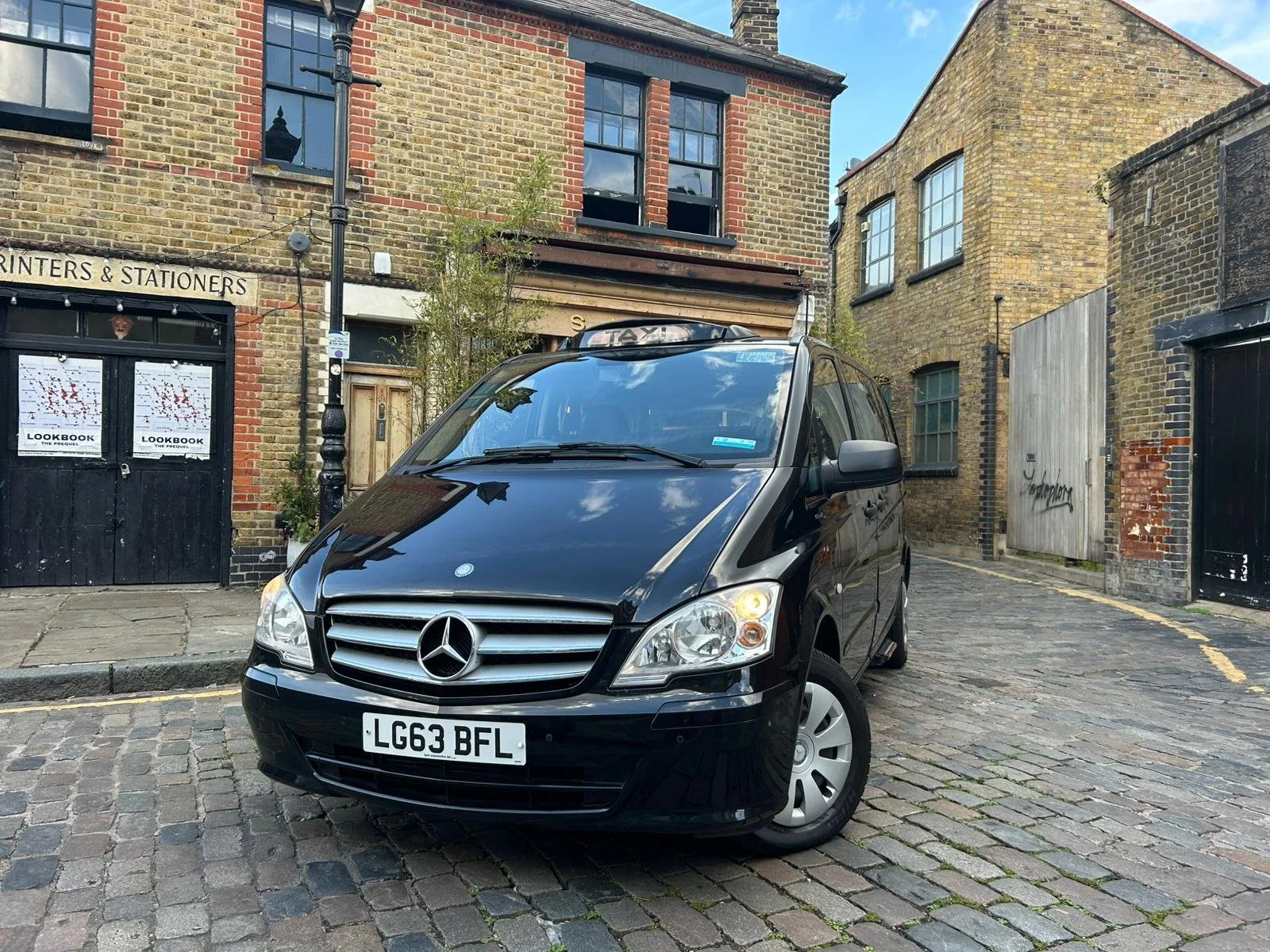 Black Mercedes-Benz taxi van parked on a cobblestone street in front of brick buildings with shop windows and graffiti on a wooden fence.