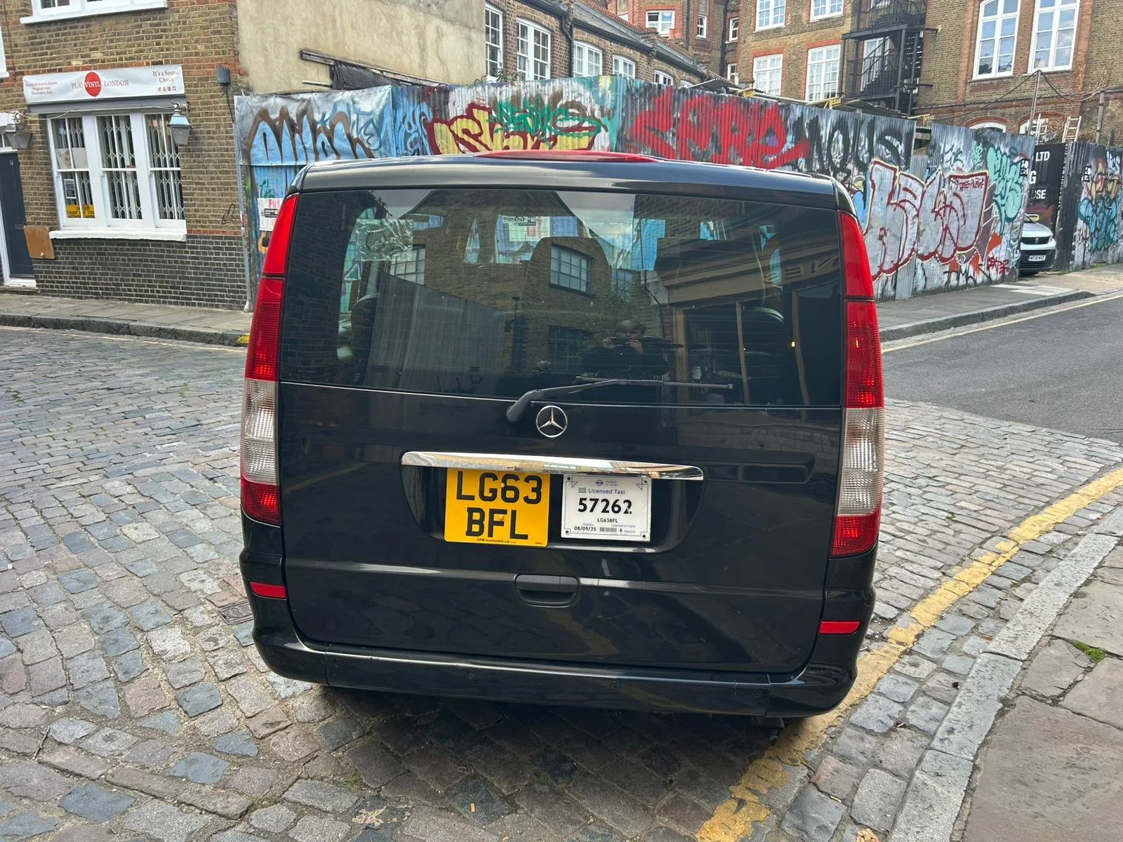 Back of a black Mercedes-Benz van parked on a cobblestone street with a yellow and white license plate that reads 'LG63 BFL' on the left and a temporary license plate on the right. There is graffiti on a wall behind the van.