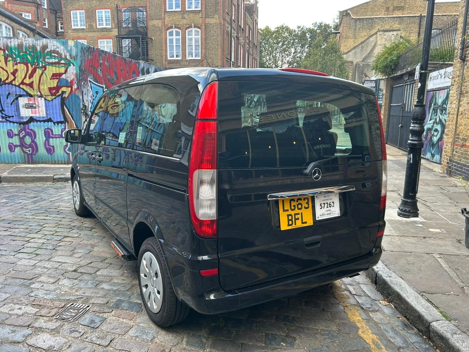Black Mercedes-Benz van is parked on a cobblestone street with colorful graffiti on a corrugated metal fence in the background. There are buildings and trees visible behind the fence.